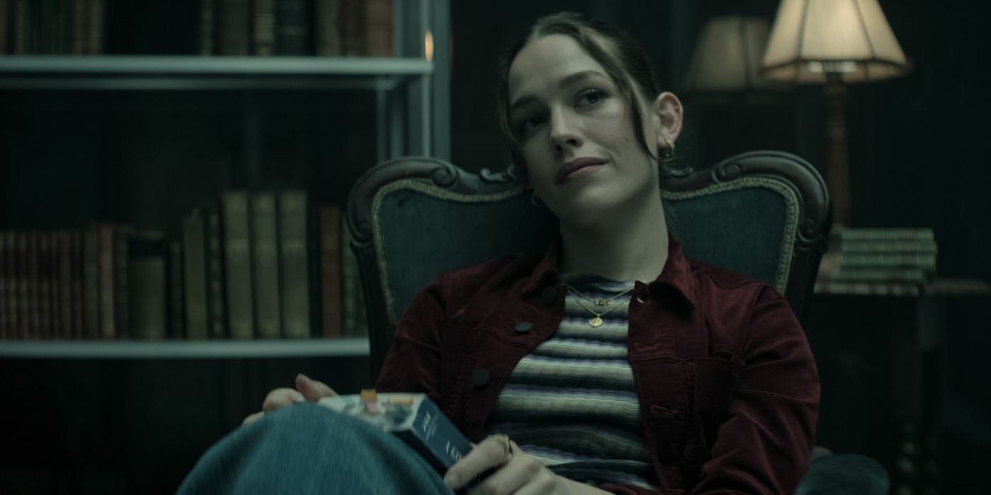 Love Quinn, played by Victoria Pedretti, sitting in a chair and holding a book in Joe's glass box during Joe's dream in You Season 4 Part II