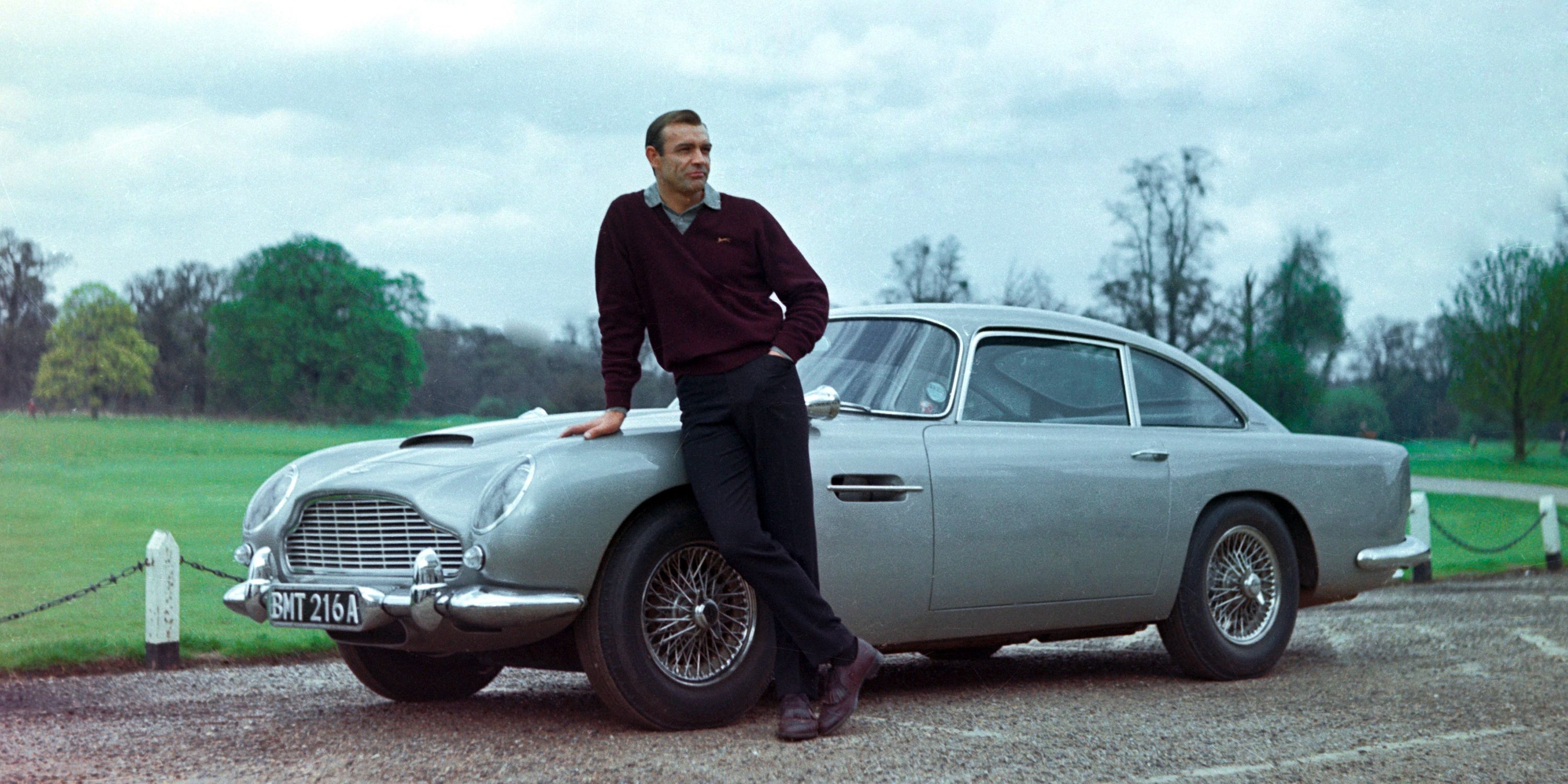 James Bond (Sean Connery) stand with his Aston Martin DB5 in a countryside driveway. 