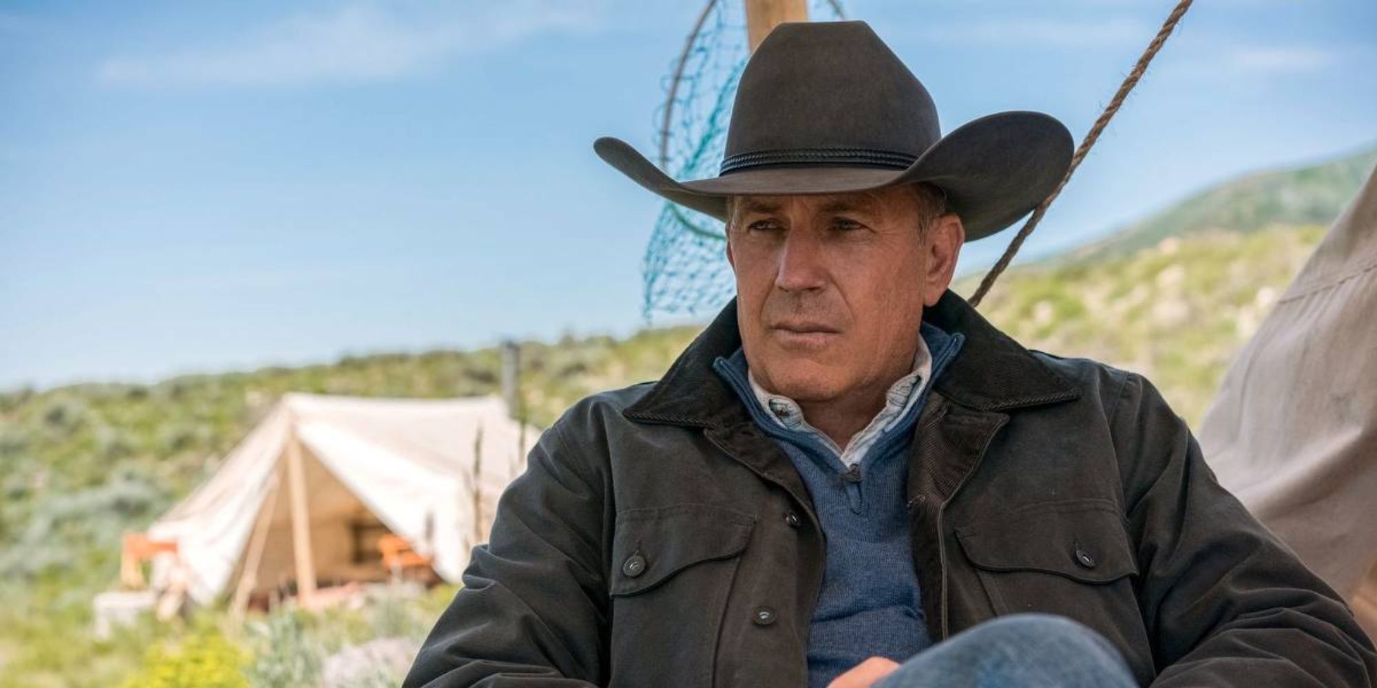 Kevin Costner in Yellowstone sitting with a cowboy hat, a tent in the background.
