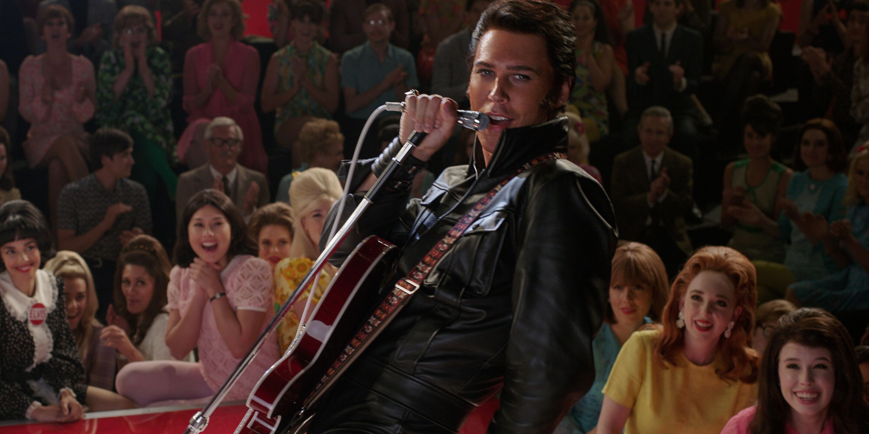 man in leather jacket singing into microphone on stage, with audience in background