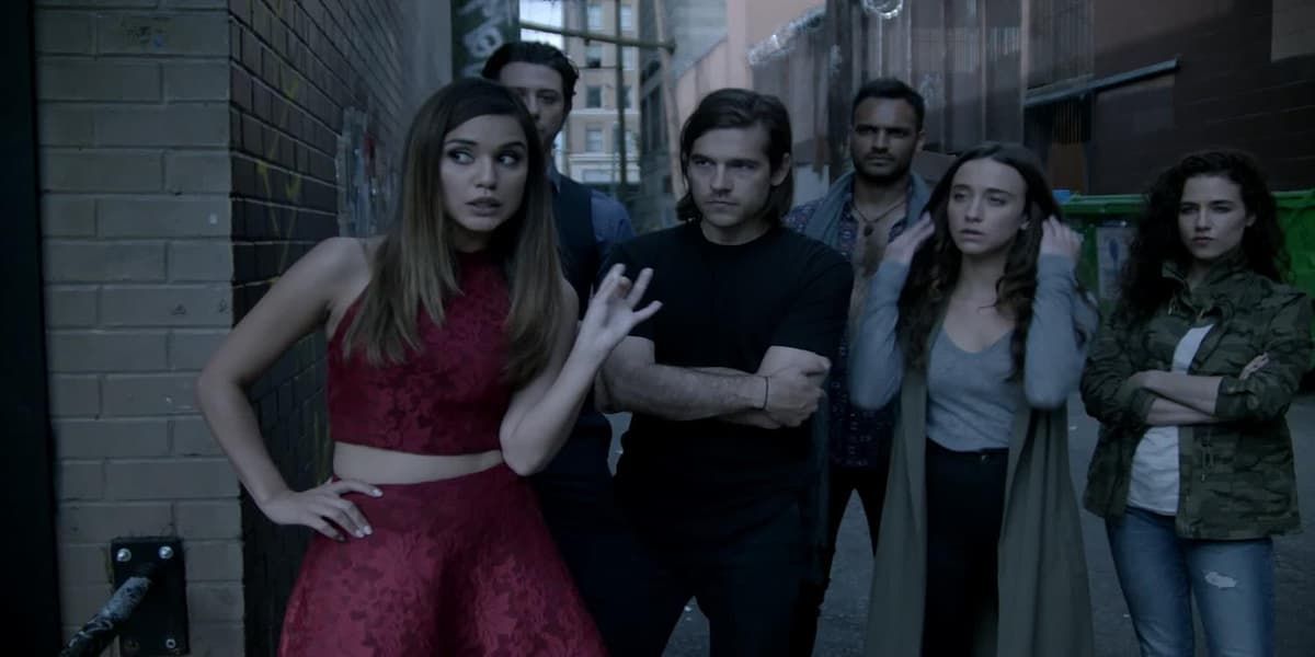 The main cast, featuring Summer Bishil, Jason Ralph, and Stella Maeve prominently stands outside of a bank
