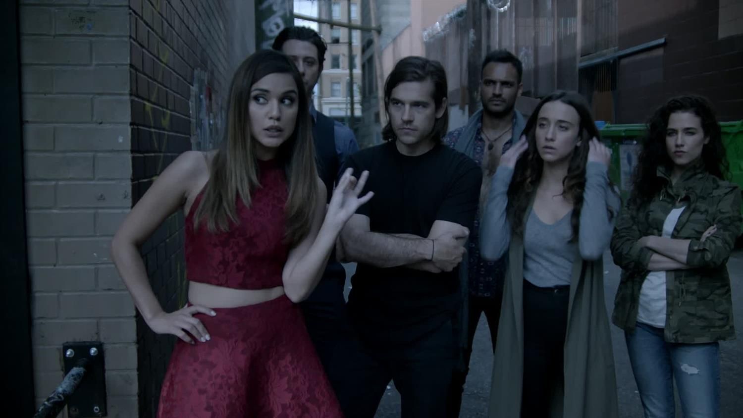 The main cast of The Magicians, featuring Summer Bishil, Jason Ralph, and Stella Maeve prominently stands outside of a bank.