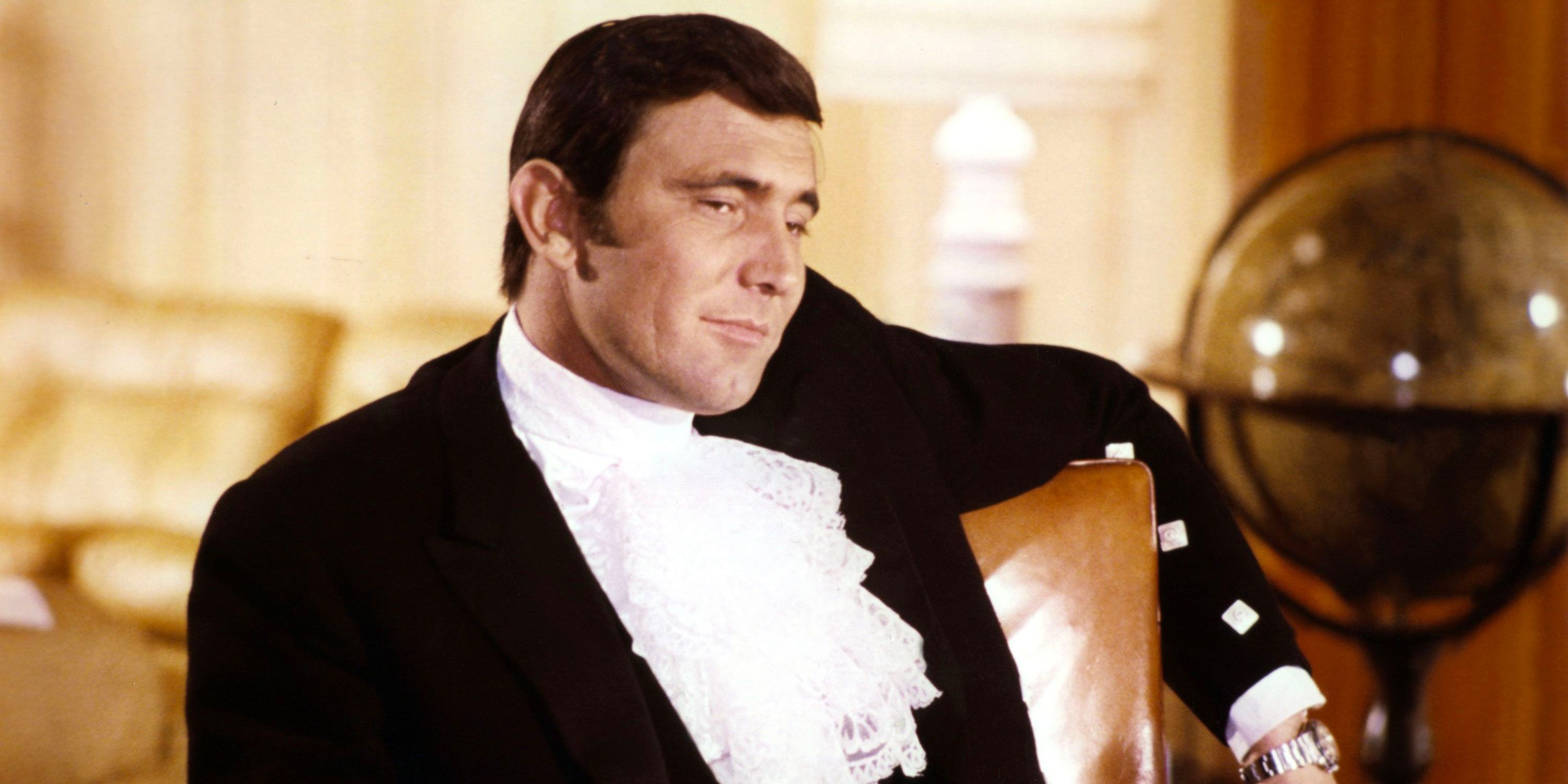 James Bond (George Lazenby) leans back in a chair while wearing a fancy suit. 