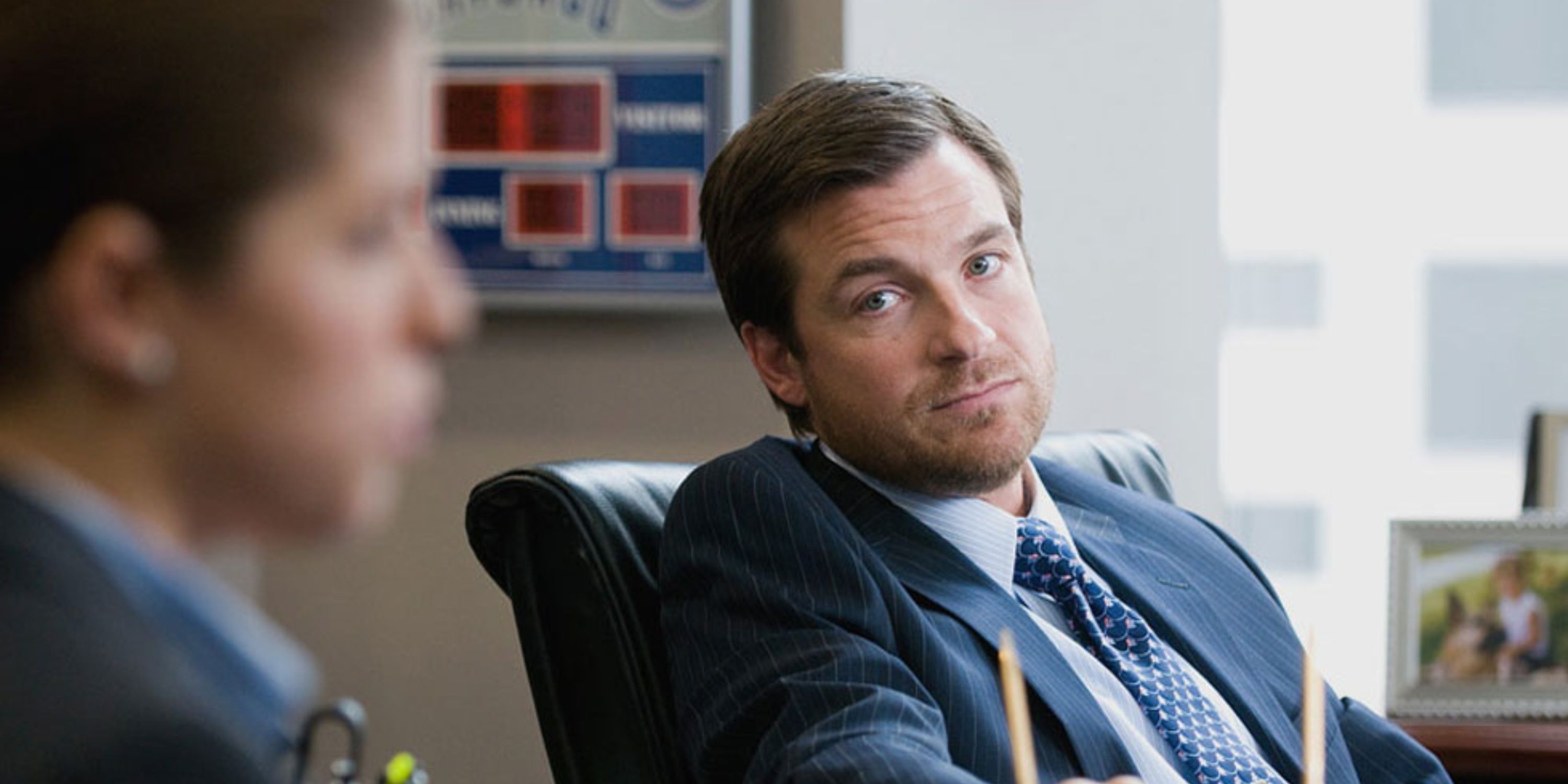 Jason Bateman as Craig Greggory leaning back on his chair and looking at another man in Up in the Air.