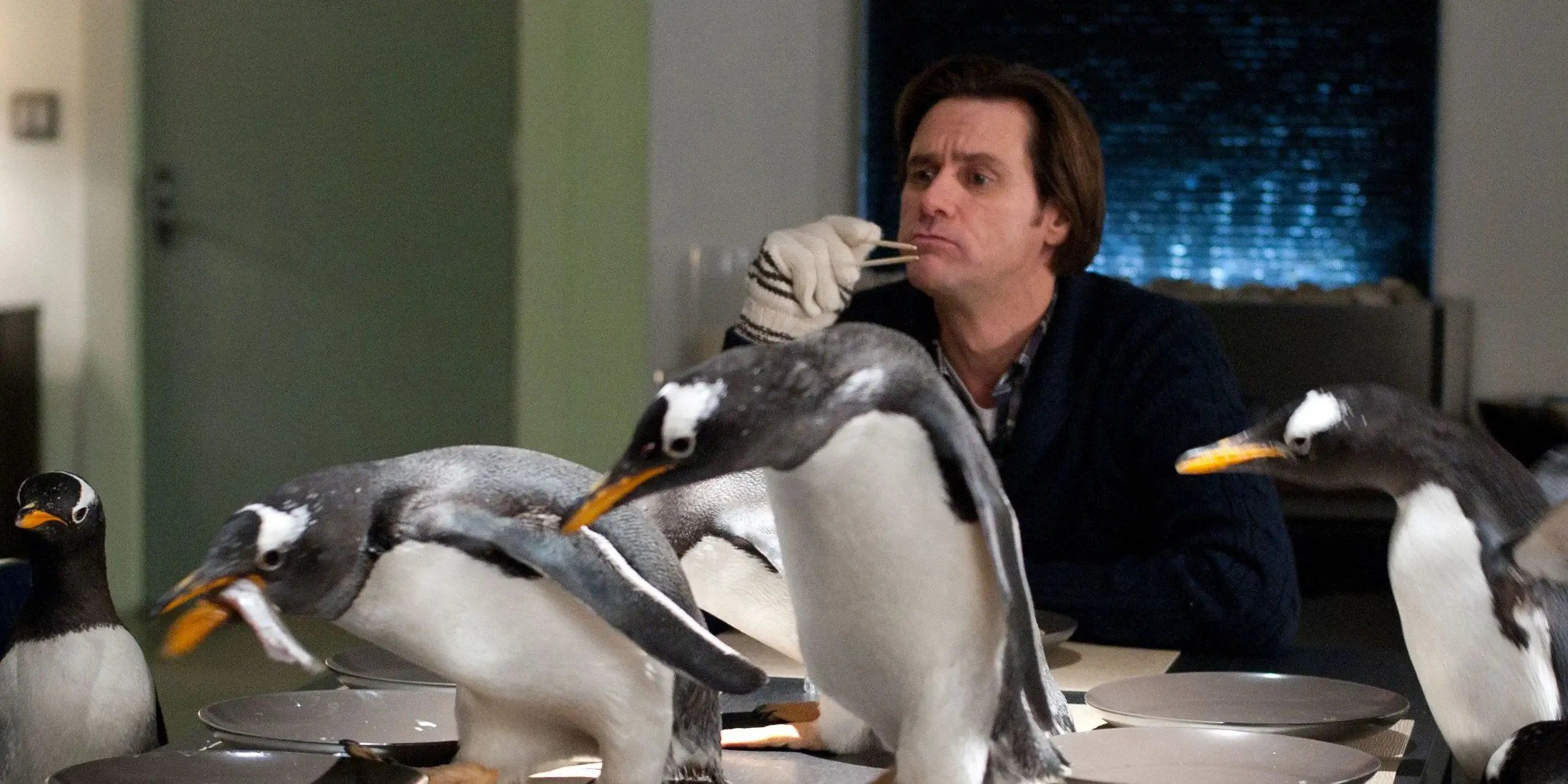 A still from the film 'Mr. Popper's Penguins' featuring Jim Carrey attempting to eat food while a group of penguins occupy the table.