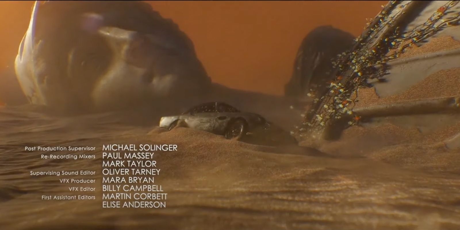 A car lies in the sand before a fallen statue during the opening credits sequence in 'No Time to Die'.