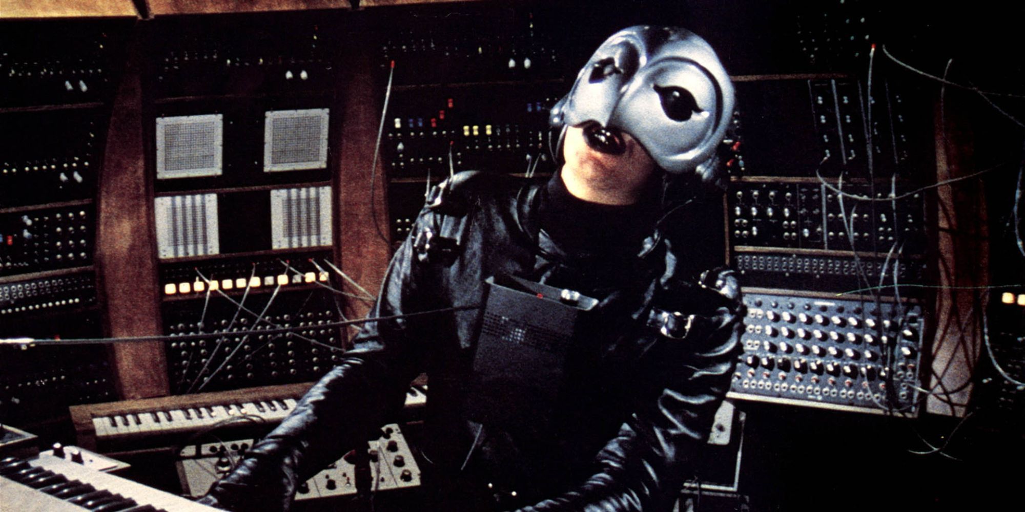 The Phantom, played by actor William Finley, sits at a large control panel strewn with wires in Phantom of the Paradise.