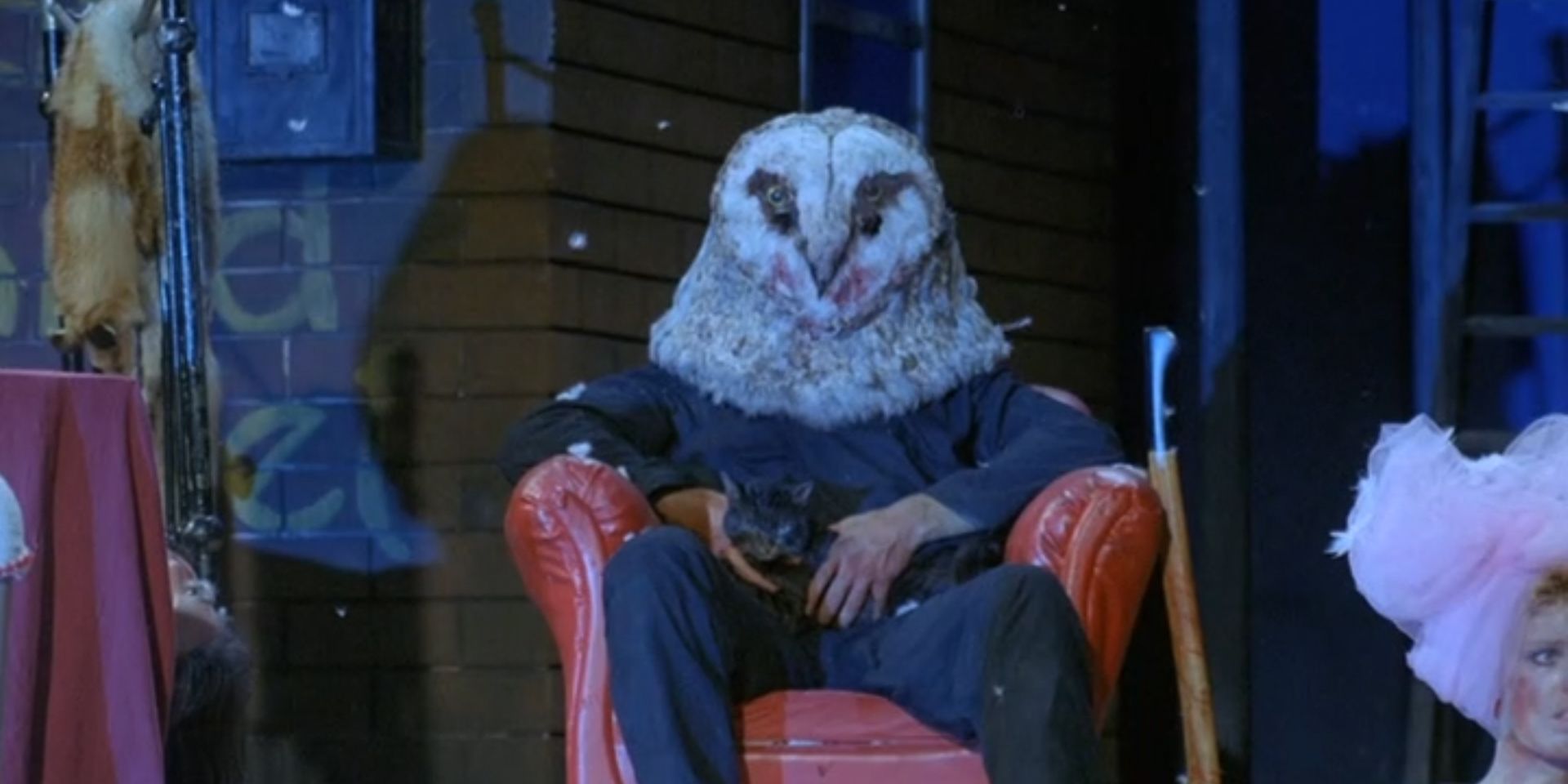 The owl-masked killer sits, petting a cat, in StageFright (1987)