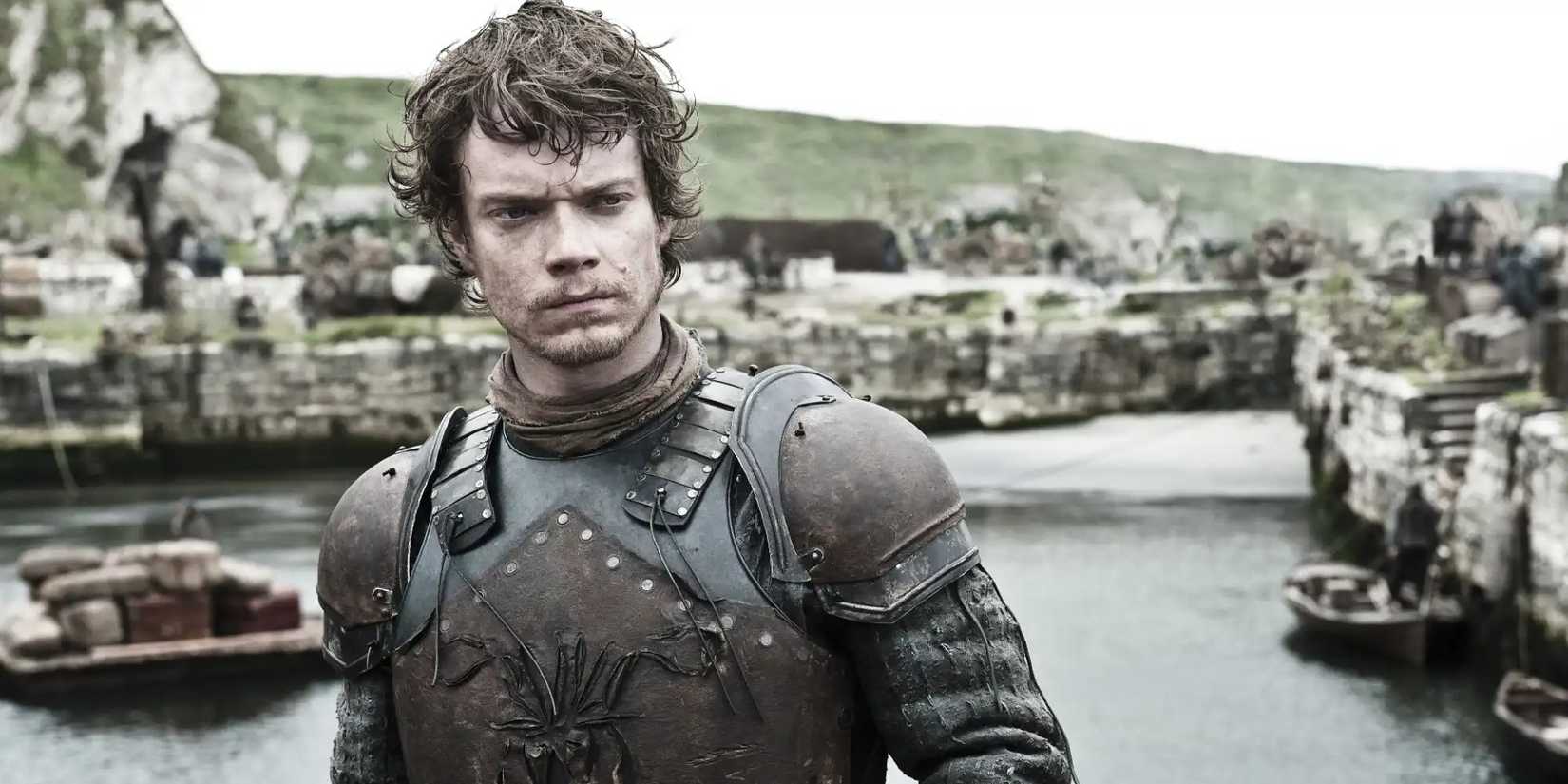 Alfie Allen as Theon Greyjoy standing on a shore looking serious in Game of Thrones.