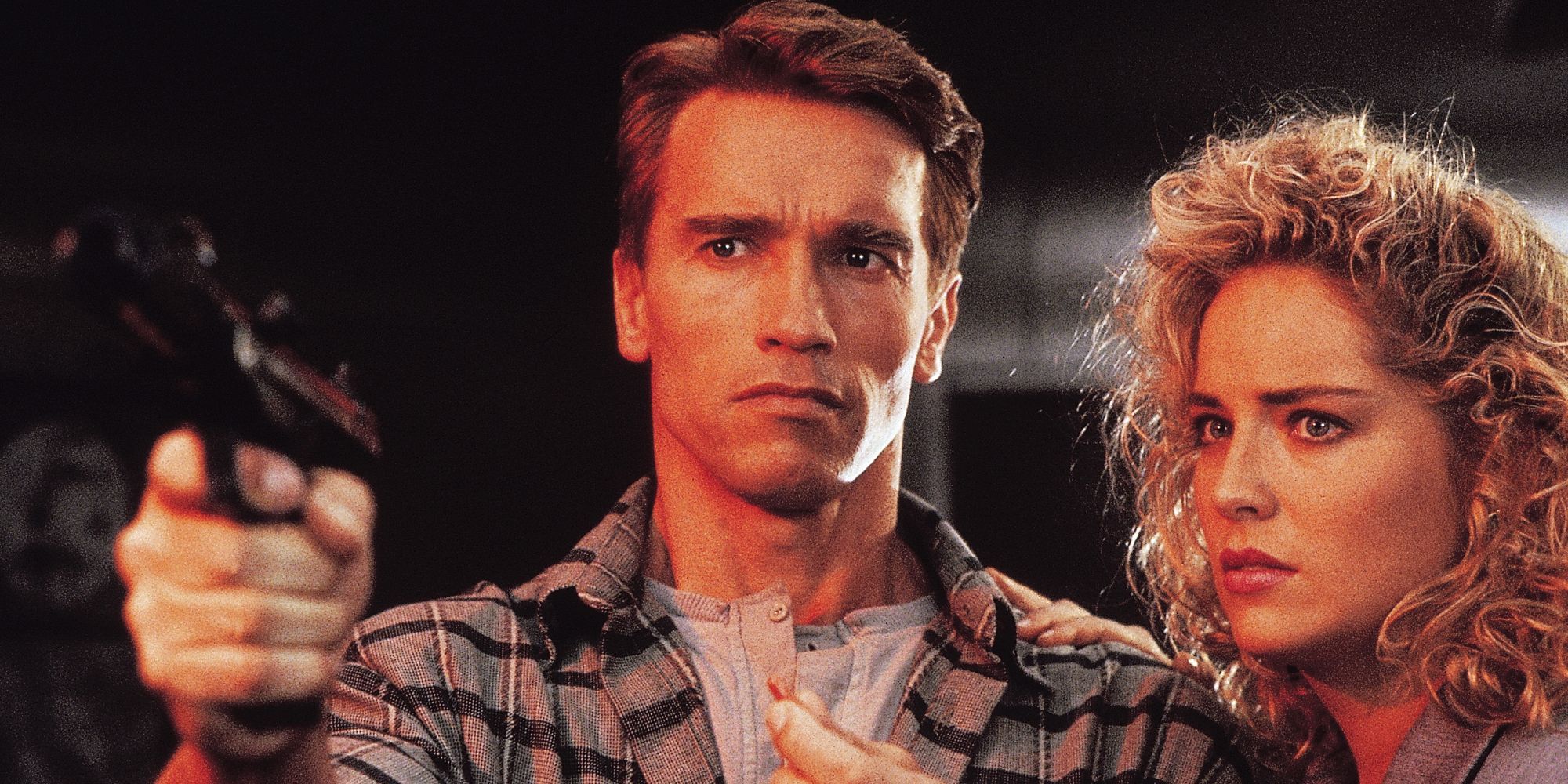 Arnold Schwarzenegger points a gun at someone as Sharon Stone holds onto him in Total Recall.