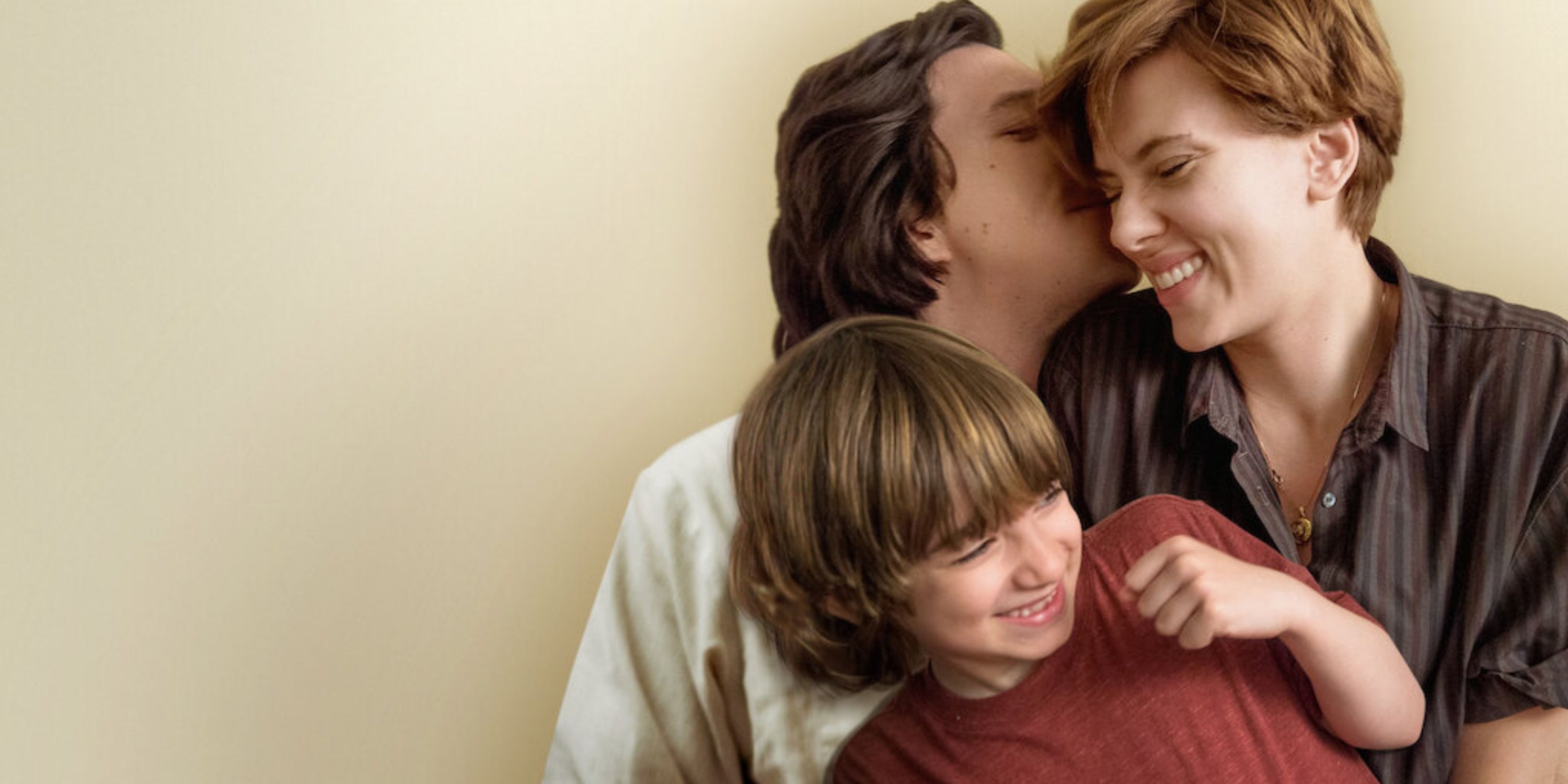 Adam Driver as Charlie Barber, kissing Scarlett Johansson as Nicole Barber, while the two are holding Azhy Robertson as Henry Barber, their son. They are infront of a beige wall.