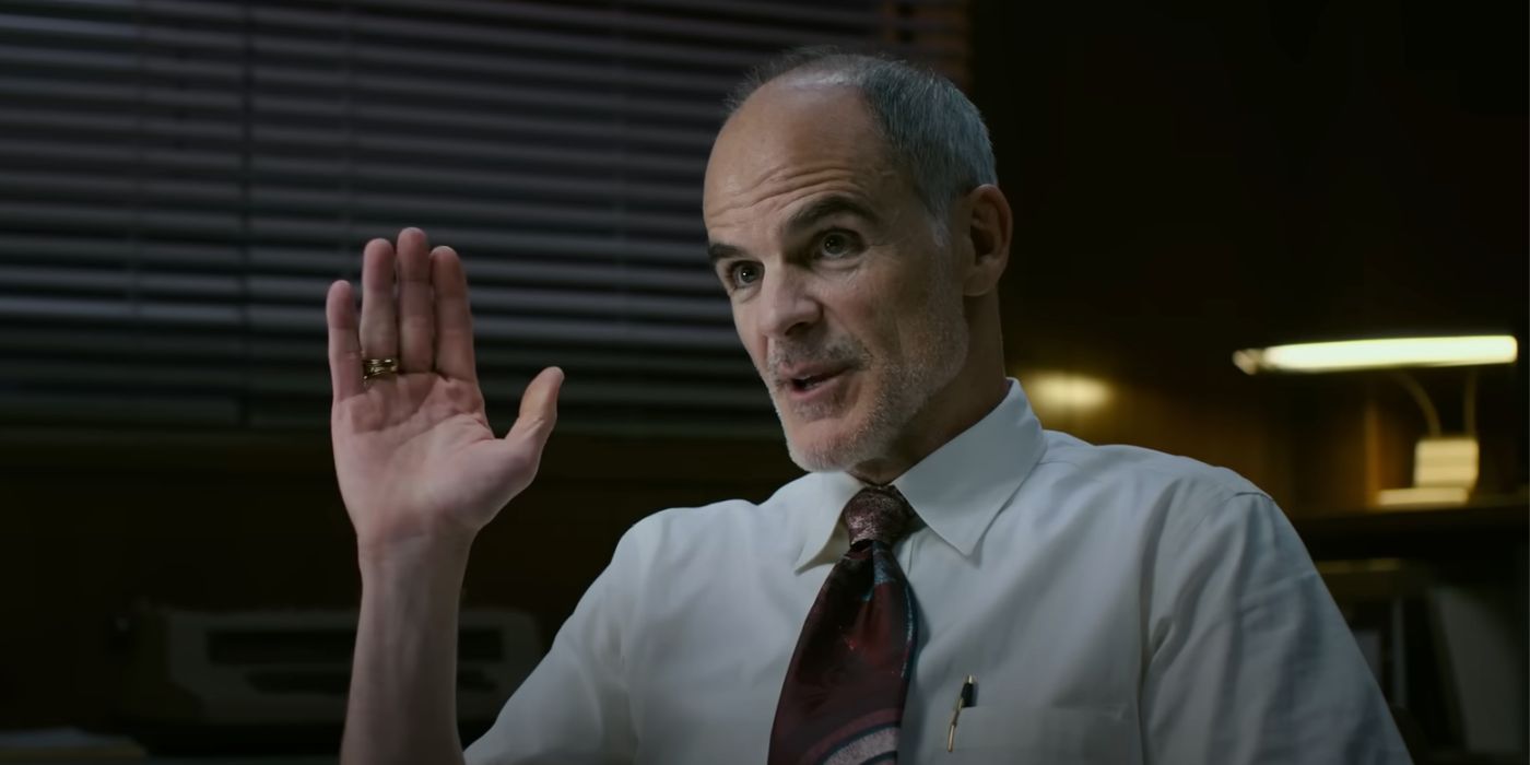 Agent Burke (Michael Kelly) wearing a white dress shirt and tie, sitting down and raising his hand in Transformers: Rise of the Beasts