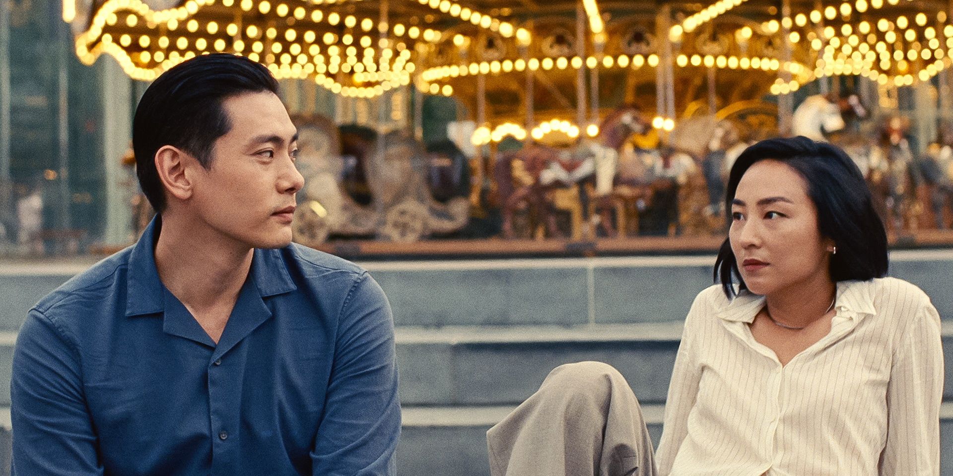 Tee Yoo and Greta Lee sitting in front of a carousel