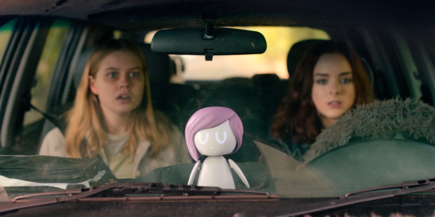 Two teenage sisters sit in a car with a robotic doll of a pop star sitting on the dashboard.