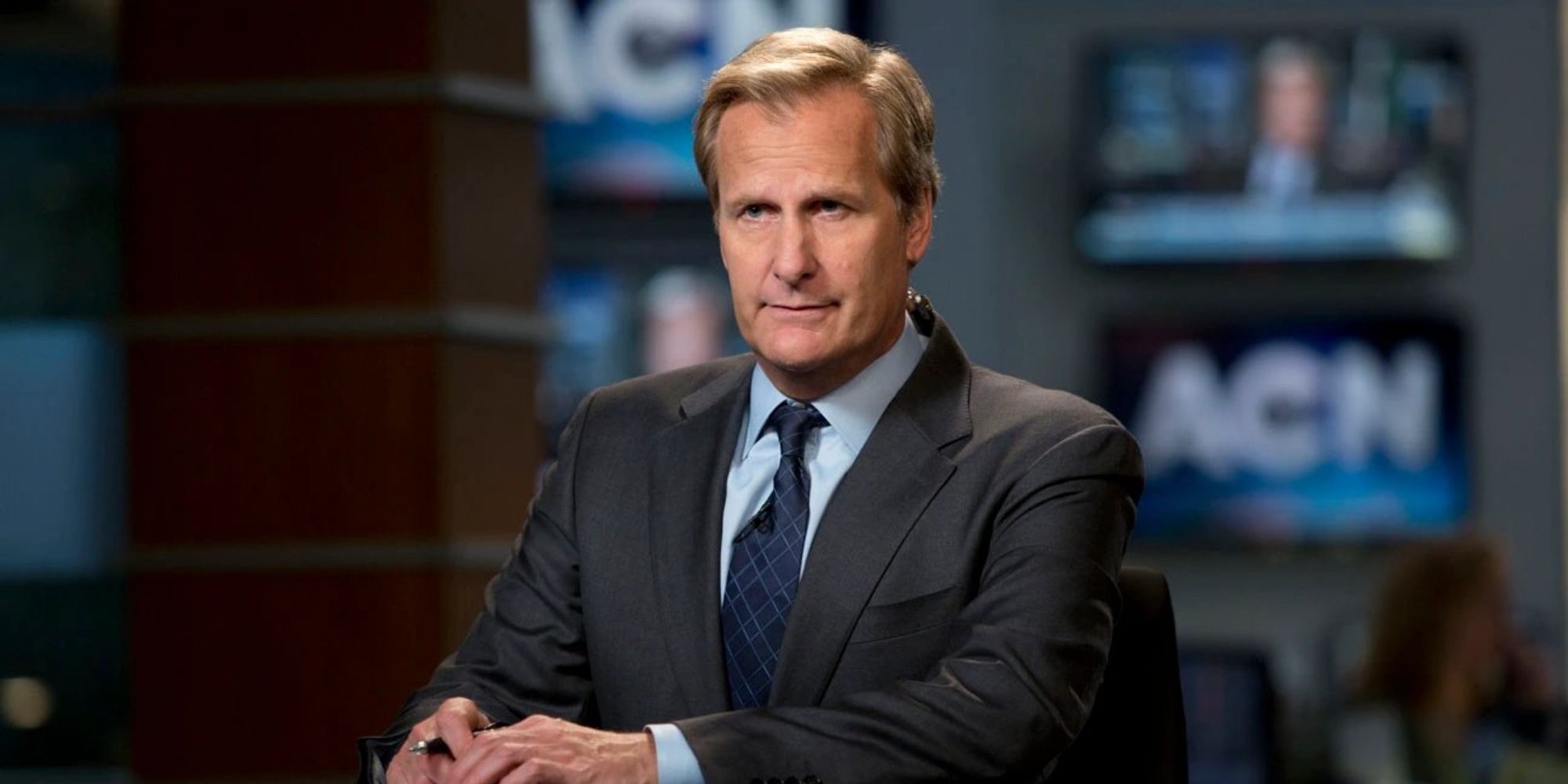 Jeff Daniels as Will McAvoy in a business suit sitting at a desk on a news program set in The Newsroom.