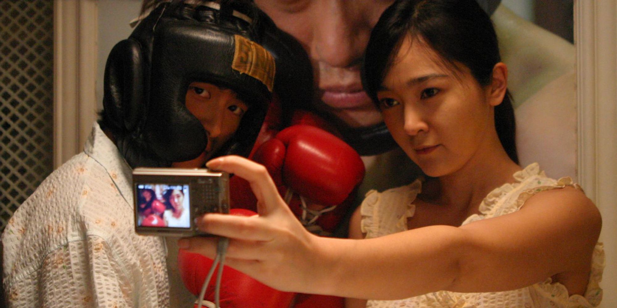 A woman taking a selfie with a digital camera with a man next to her, who's wearing a boxing helmet and gloves