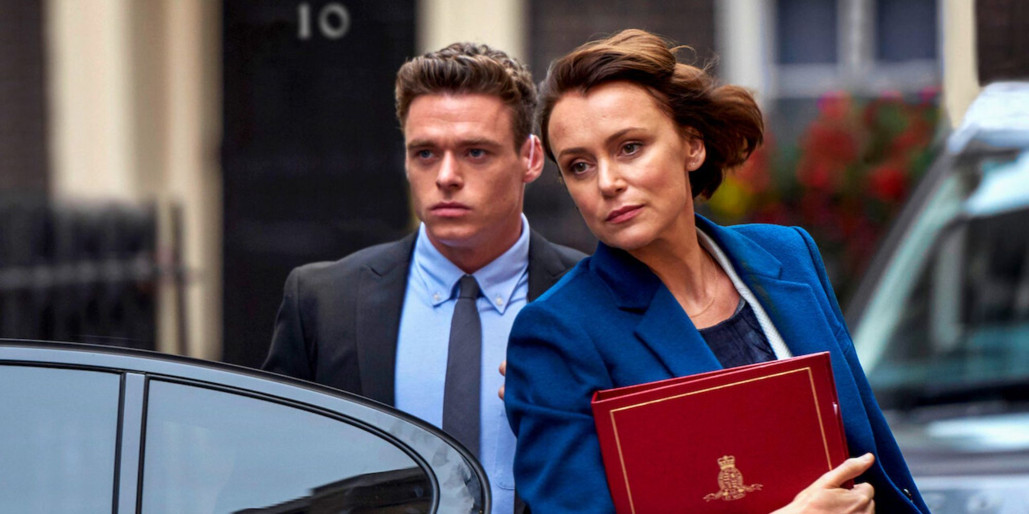 A man in a suit escorts a woman with a binder into a car in a scene from Bodyguard.