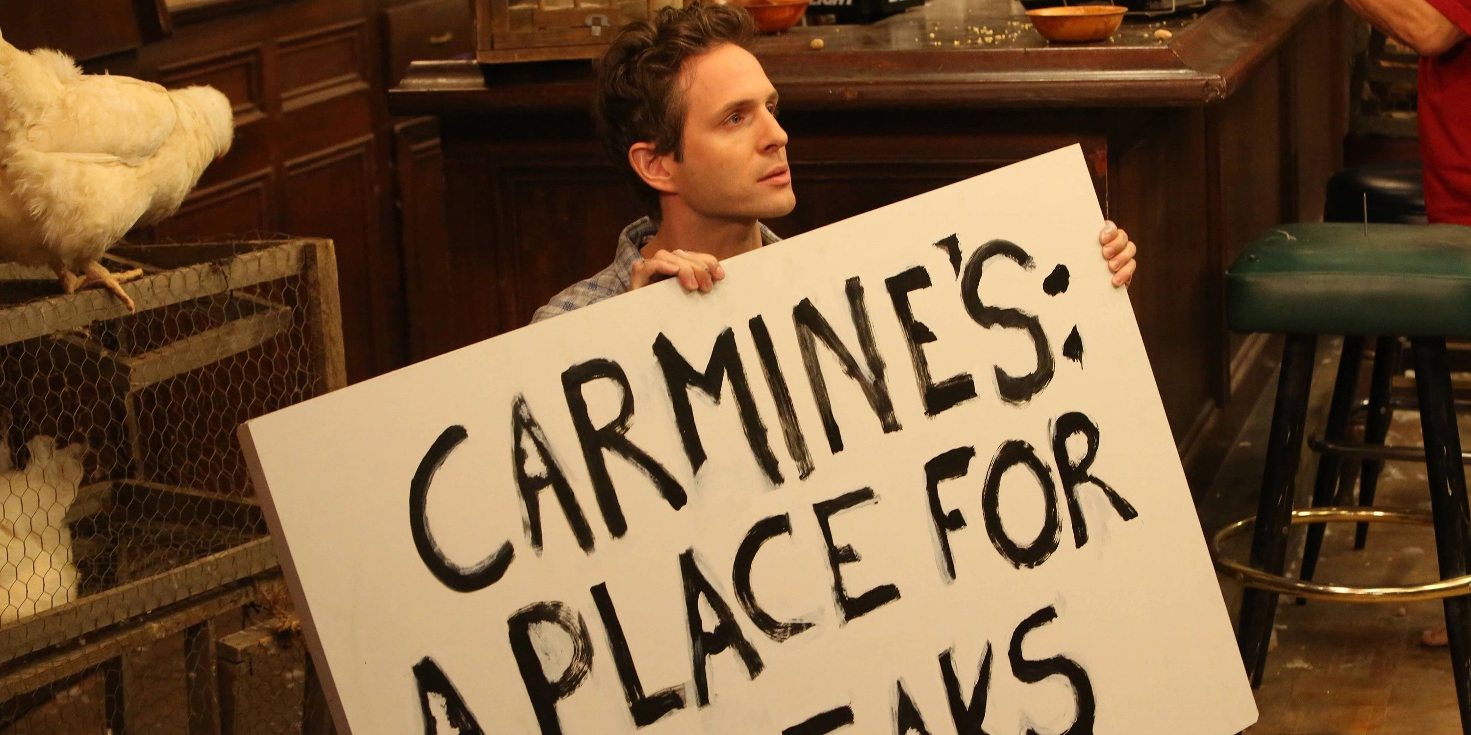 Still from 'It's Always Sunny in Philadelphia': Dennis (Glenn Howerton) sits on the floor with a sign reading "CARMINE'S: A PLACE FOR STEAKS" next to a cage of chickens."