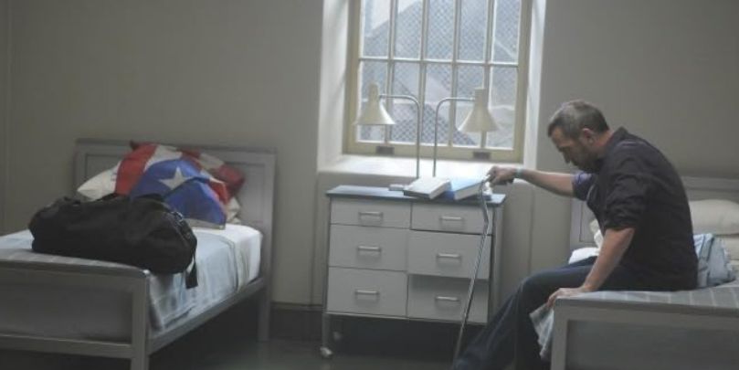 Hugh Laurie as Dr. Gregory House sitting on a bed in a psych ward room in 'House, M.D.'