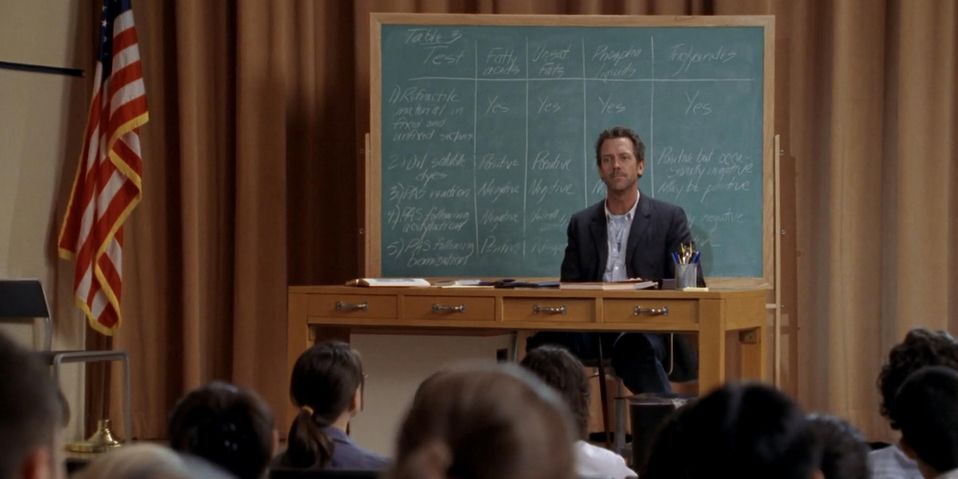 Hugh Laurie as Dr. Gregory House in 'House, M.D.', he is sitting in a desk in front of a chalkboard giving a lecture
