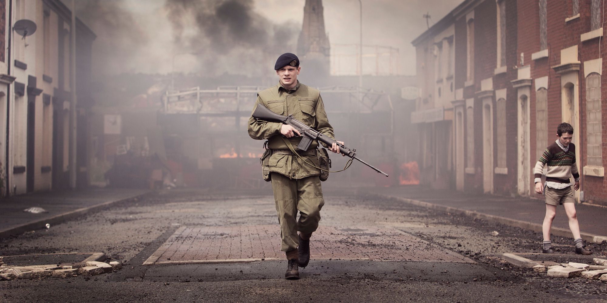 Jack O'Connell walking down a destroyed street with a firearm in 