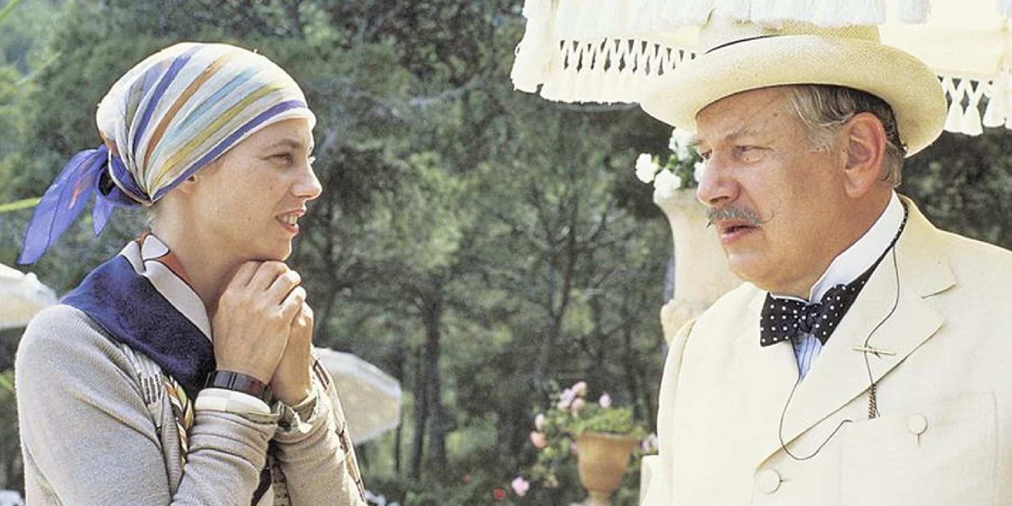 Poirot, who is holding a white umbrella, talks with a young woman outside.