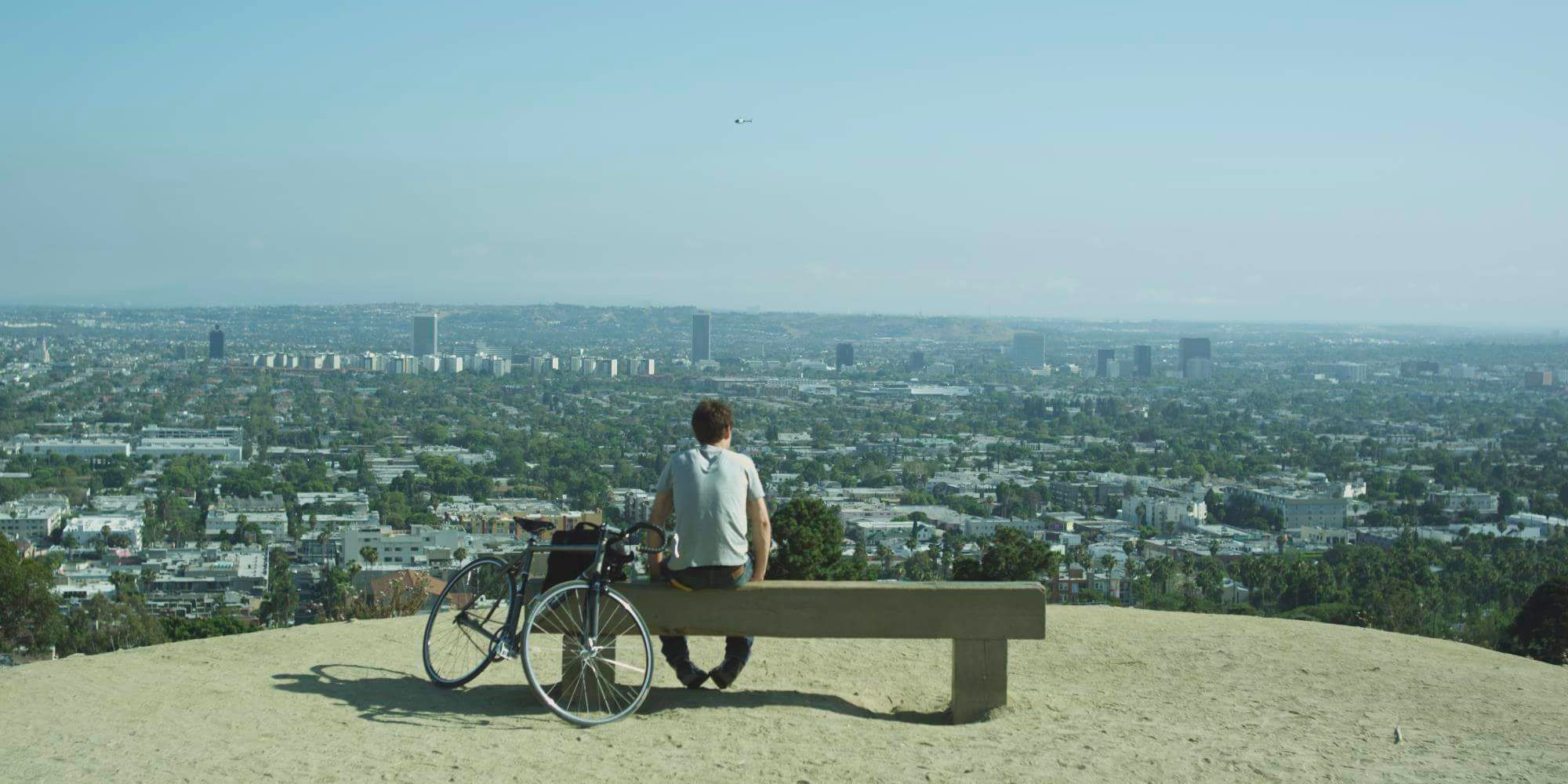 A man sits on a bench on a sunny day looking out over the city of Los Angeles with his bike.