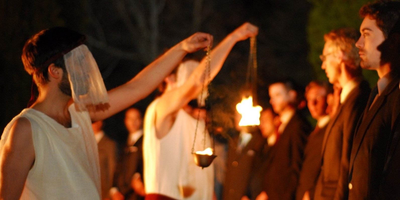 Image from 'The Conspiracy' of two men in veils and robes holding flames up to a line of men.