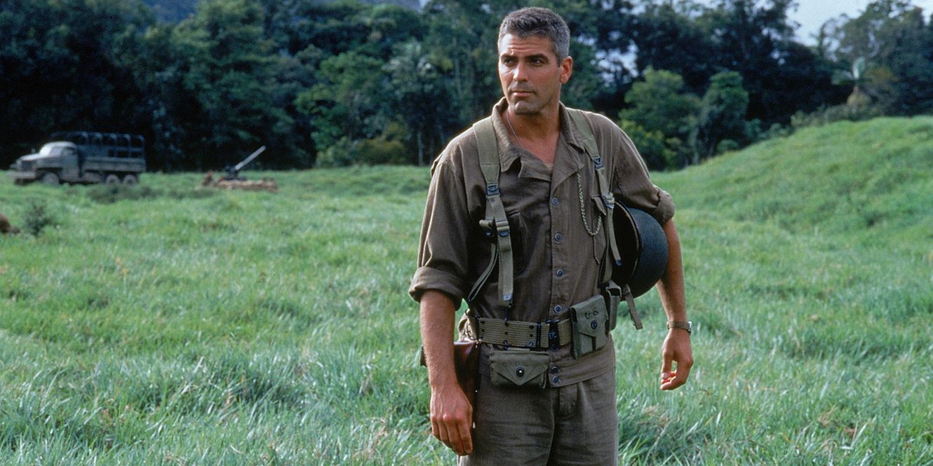 A WWII soldier stands in a green field with his helmet tucked under his arm. 