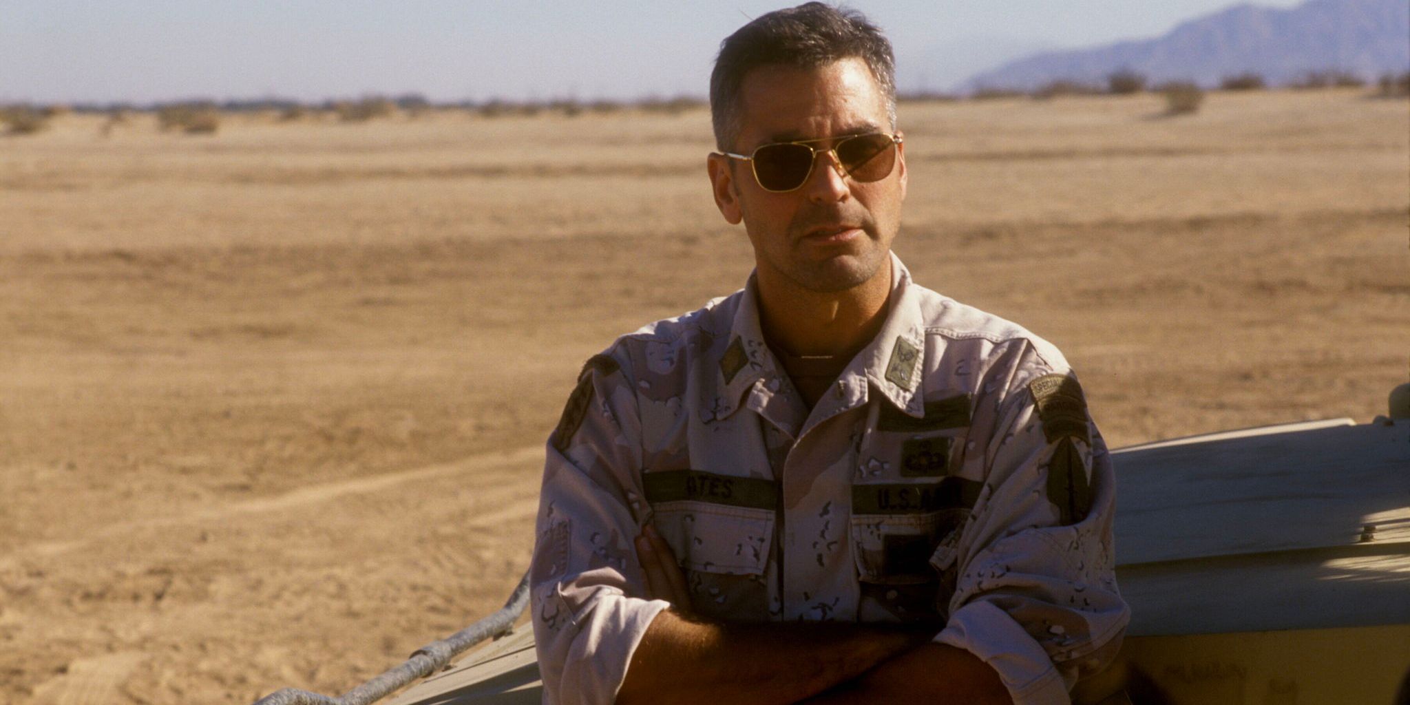 An American army man stands against a military vehicle with a barren desert behind him.