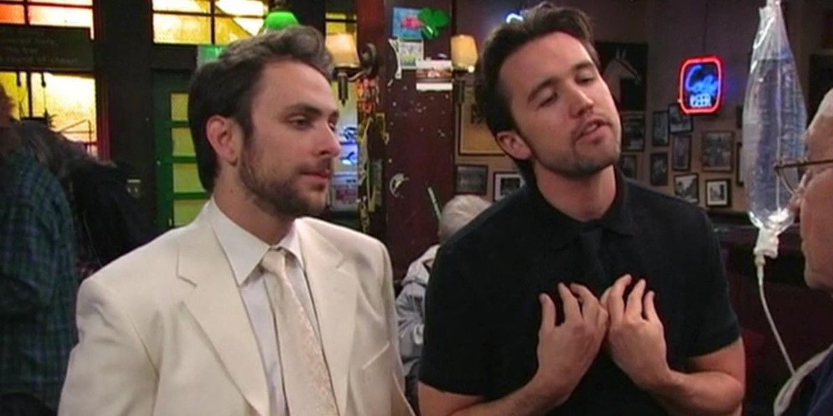 Still from 'It's Always Sunny in Philadelphia': Charlie wears a white suit and stands beside Mac, who is wearing black and talking to an elderly man.