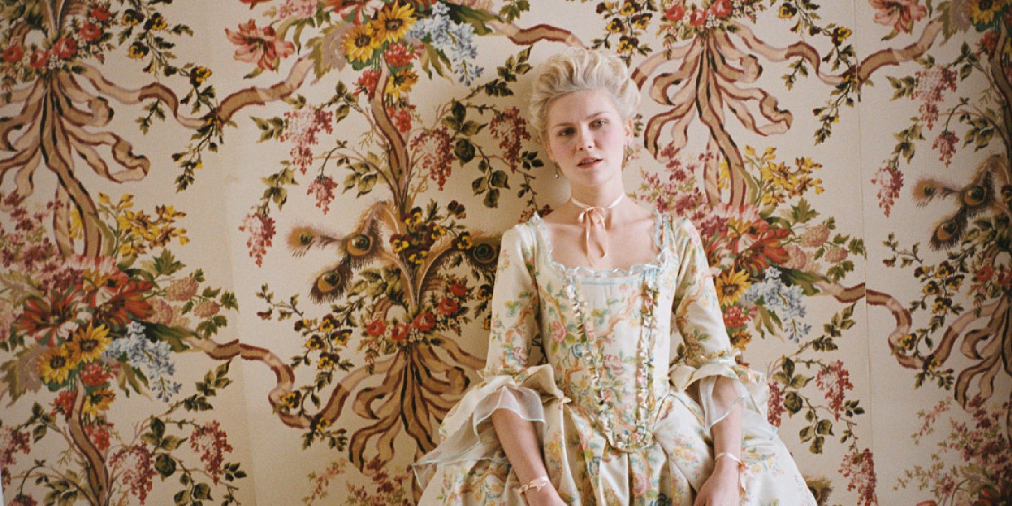 Kirsten Dunst as Marie Antoinette stands next to a wall covered in floral wallpaper.