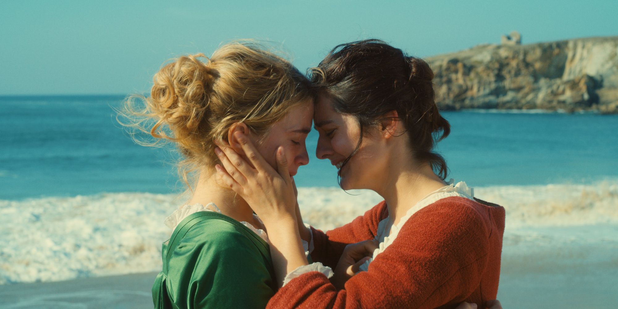 Two women, one in a green dress, the other in red, embrace each other on the shores of a pristine beach.