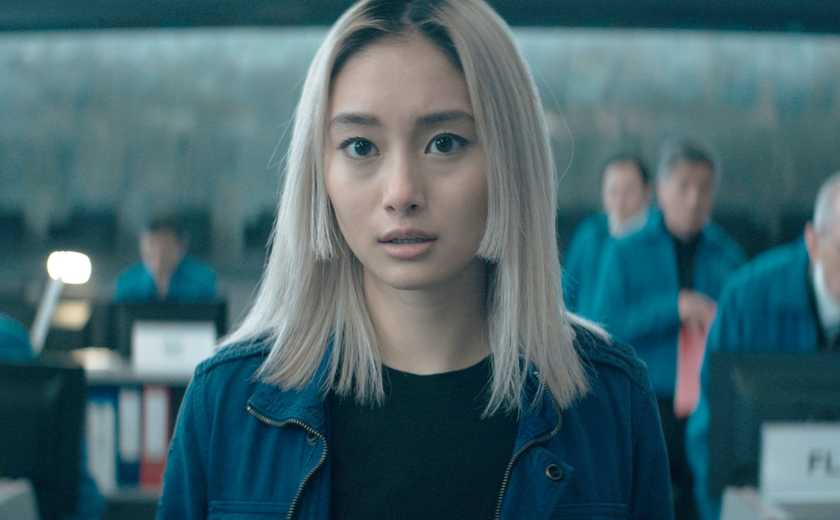 Shiolo Kutsana looks at the camera; several people sit at desks behind her, in 'Invasion'