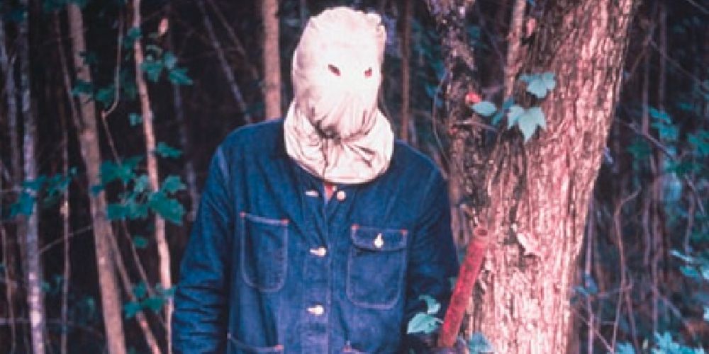A hooded murderer in a denim jacket stands in the woods with a pipe in his hand.