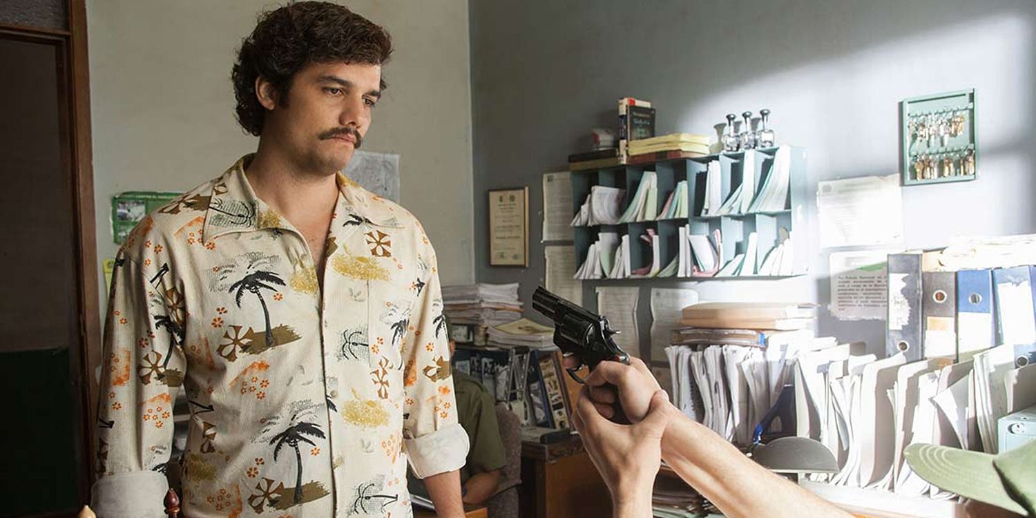 Wagner Moura as Pablo Escobar, having a gun pointed at him in an office