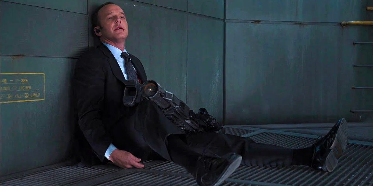 Agent Phil Coulson sits motionless on the ground with a large gun in his lap as blood trickles from his mouth.