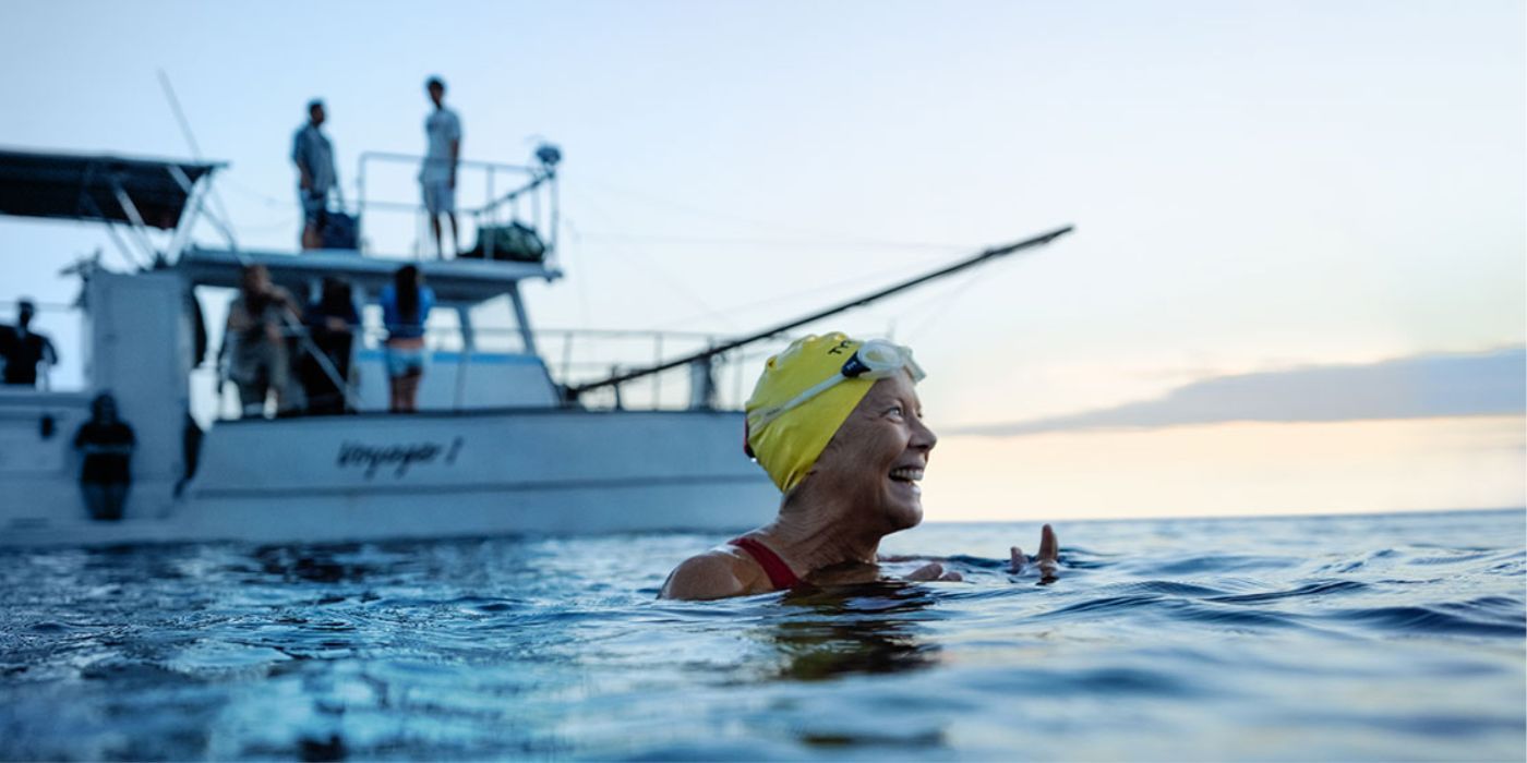 Annette Bening as Diana Nyad, wearing a swim cap and smiling in the water by a boat in Nyad