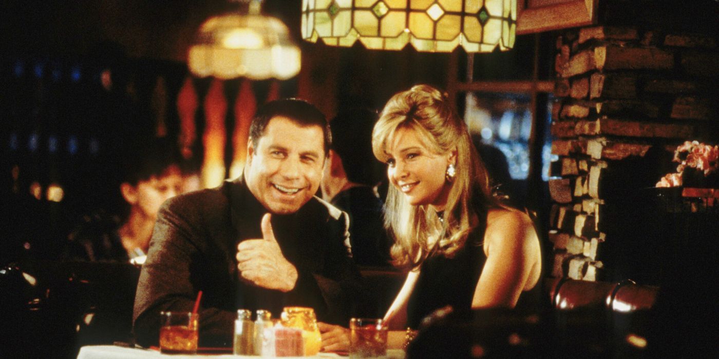 Russ (John Travolta) sitting next to Crystal (Lisa Kudrow) at dinner, smiling and giving someone a thumbs up in Lucky Numbers