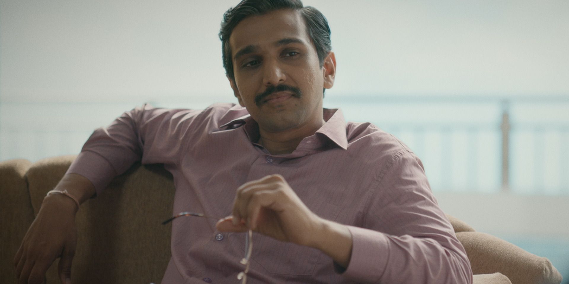 Pratik Gandhi as Harshad Mehta sitting on a couch, holding his glasses