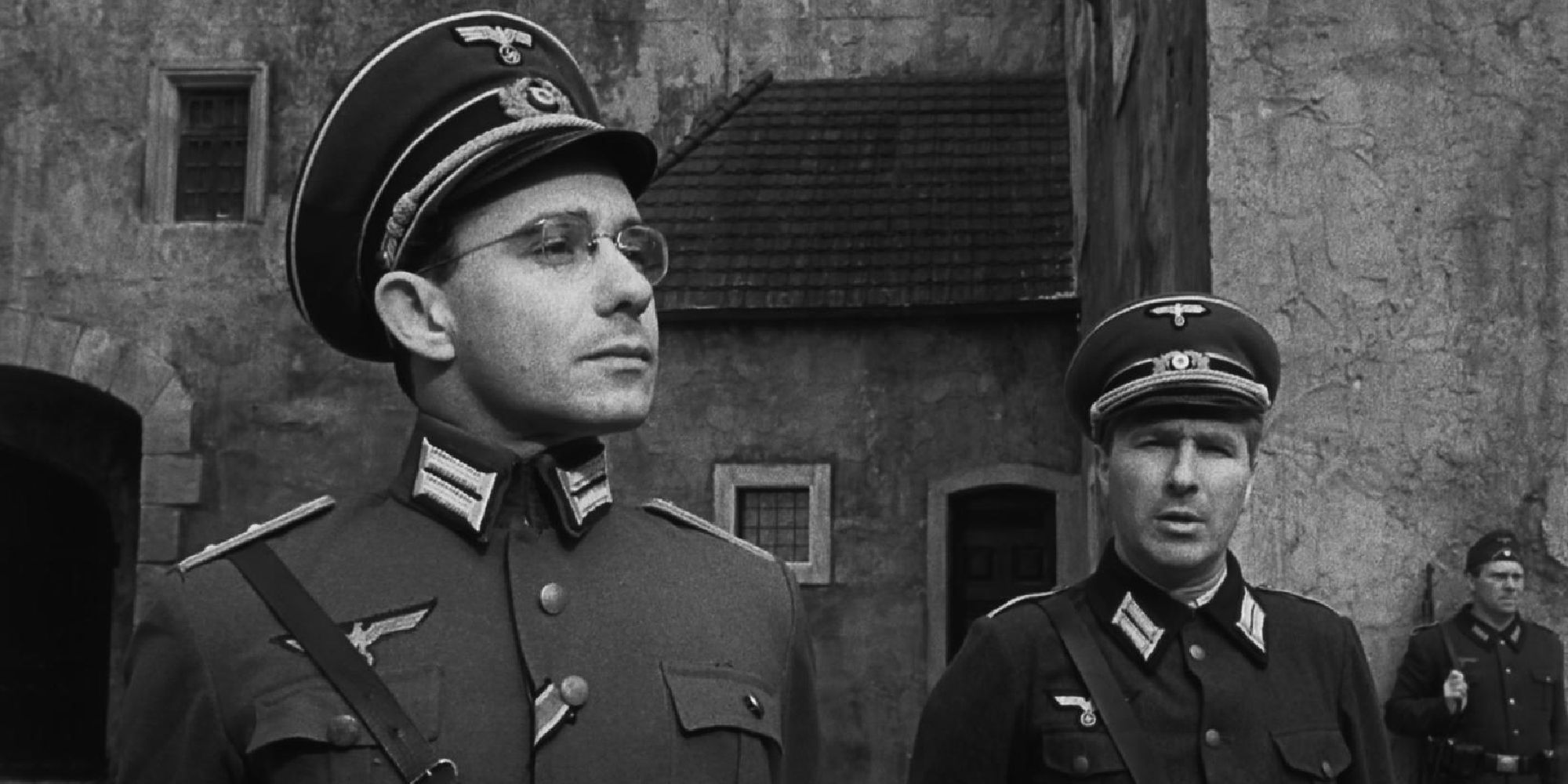 Three men in military uniforms stand outside a larger stone house in The Colditz Story.