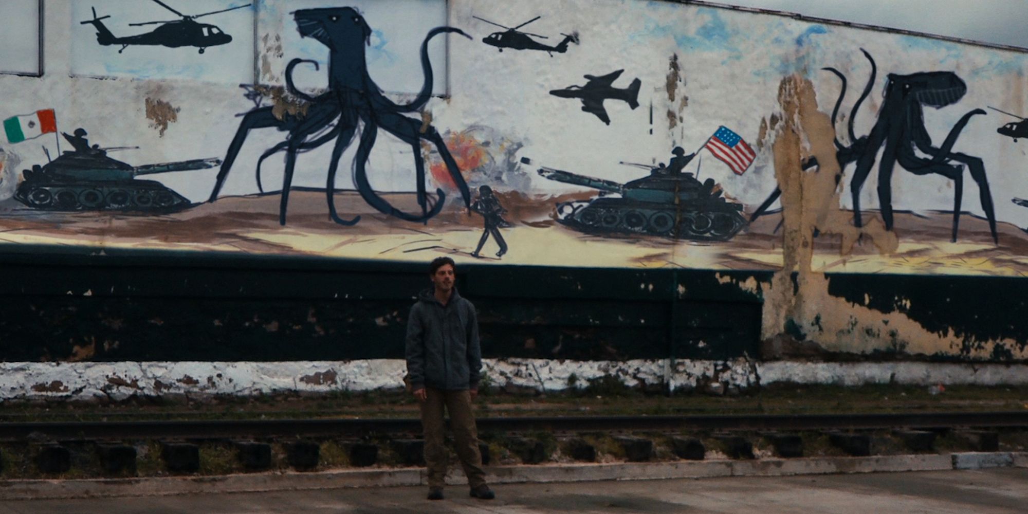 Still image of a man on traintracks with a mural behind him 