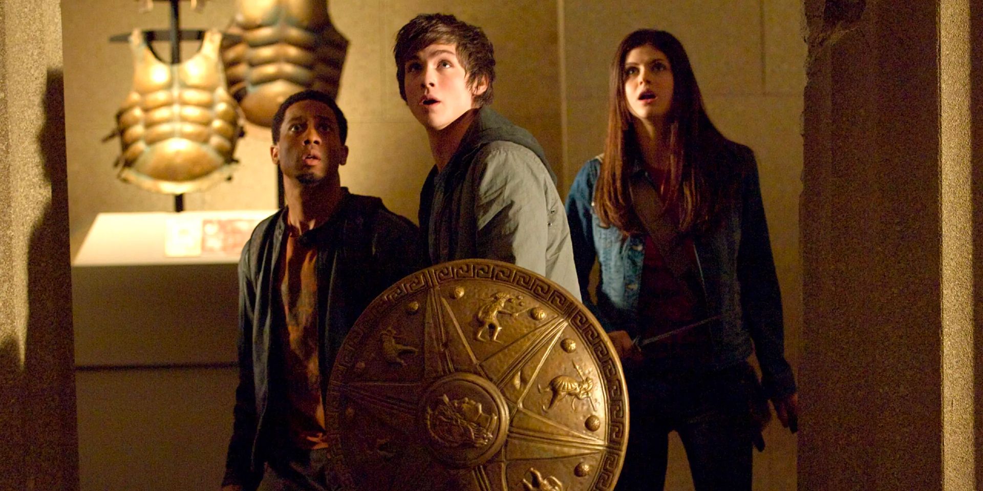 A teenage boy stands in a museum with a shield with two of his friends behind him looking up at something.