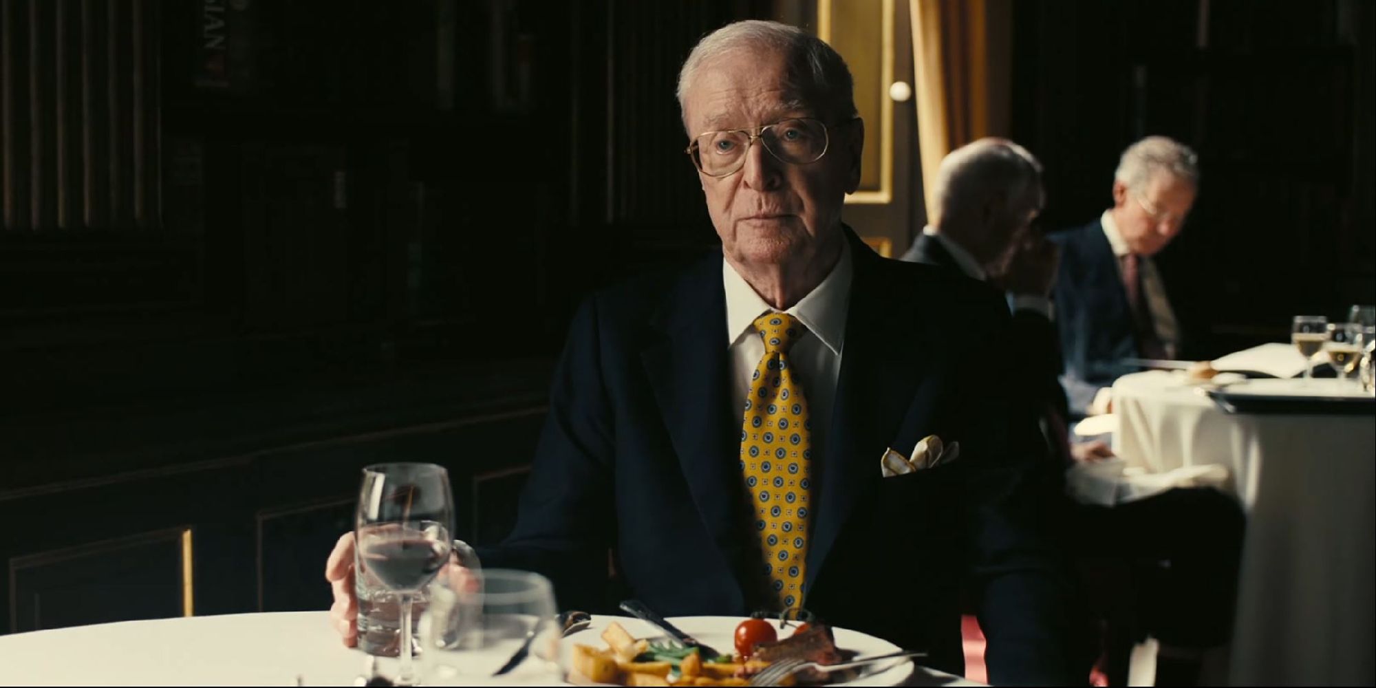 Michael Caine as Michael Crosby, sitting with a plate of food in front of him and looking solemn in Tenet