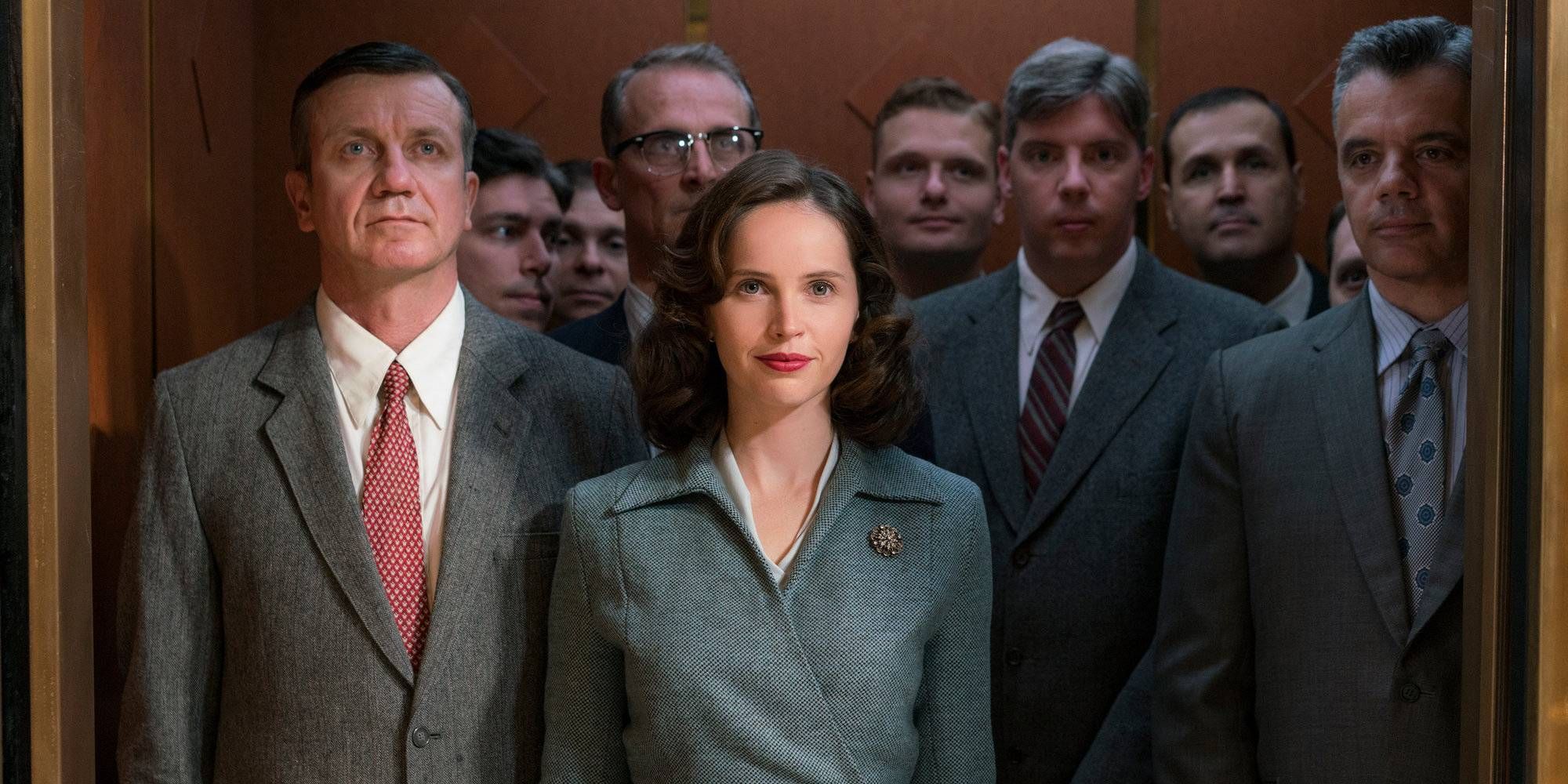 Felicity Jones as Ruth Bader Ginsburg standing on an elevator surrounded by men and smiling in On the Basis of Sex.