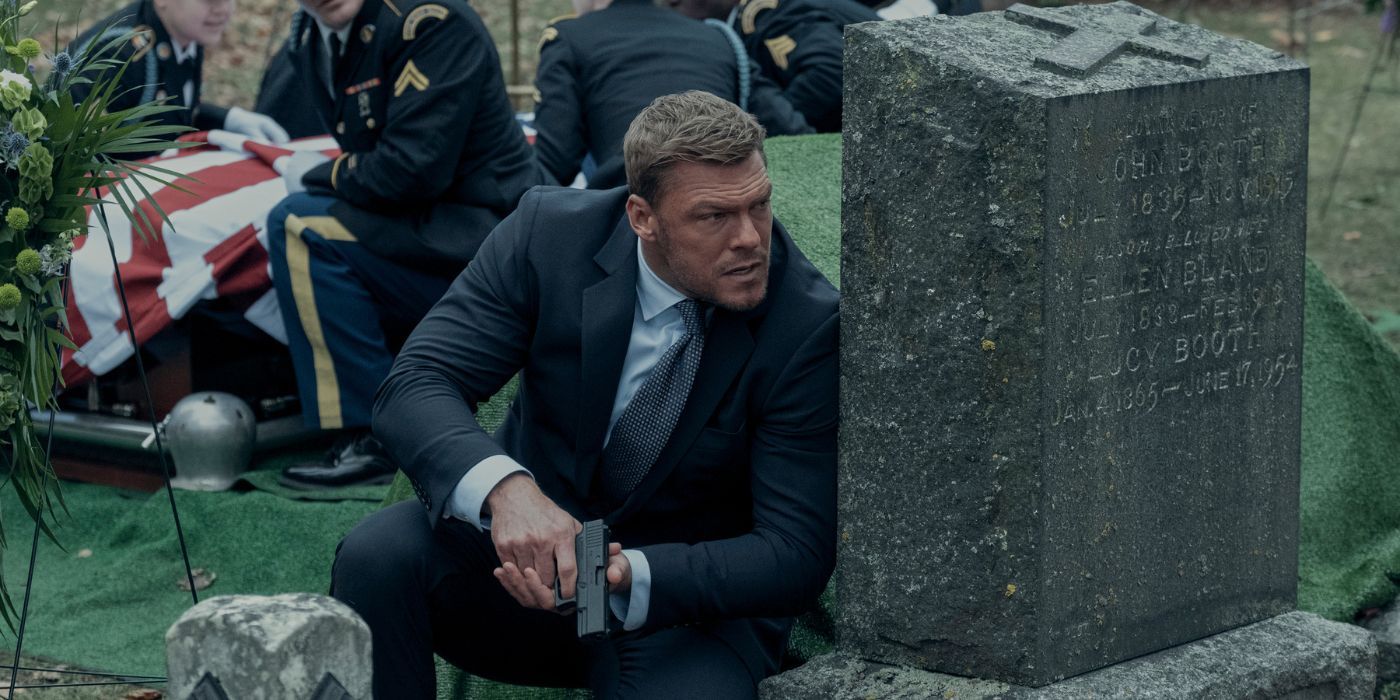 Alan Ritchson as Jack Reacher clutching a gun behind a tombstone in Reacher Season 2