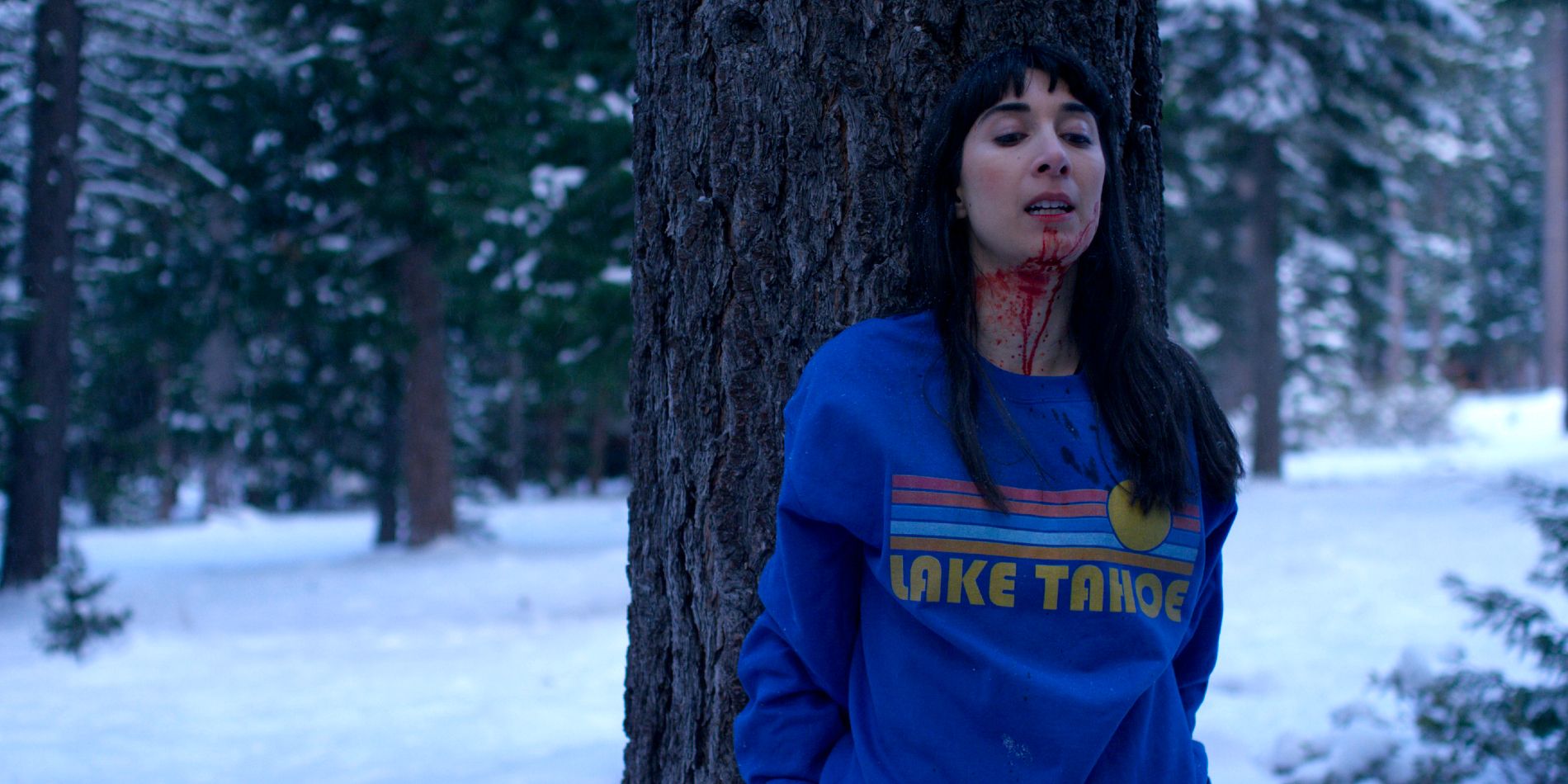 A woman with a bloodied face hides behind a tree in a snowy forest. 