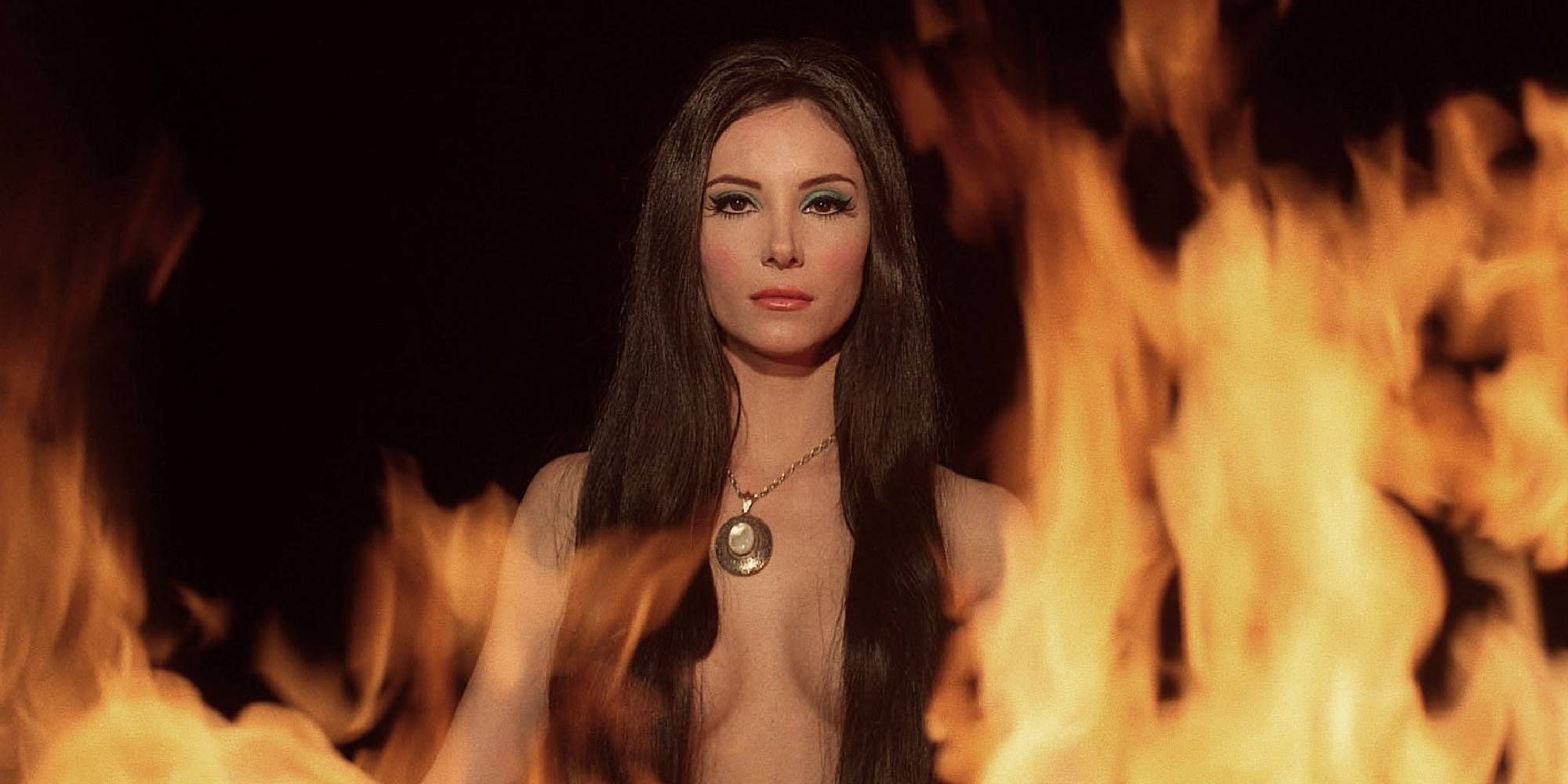 Samantha Robinson looking at the camera in The Love Witch with fire flames around her.