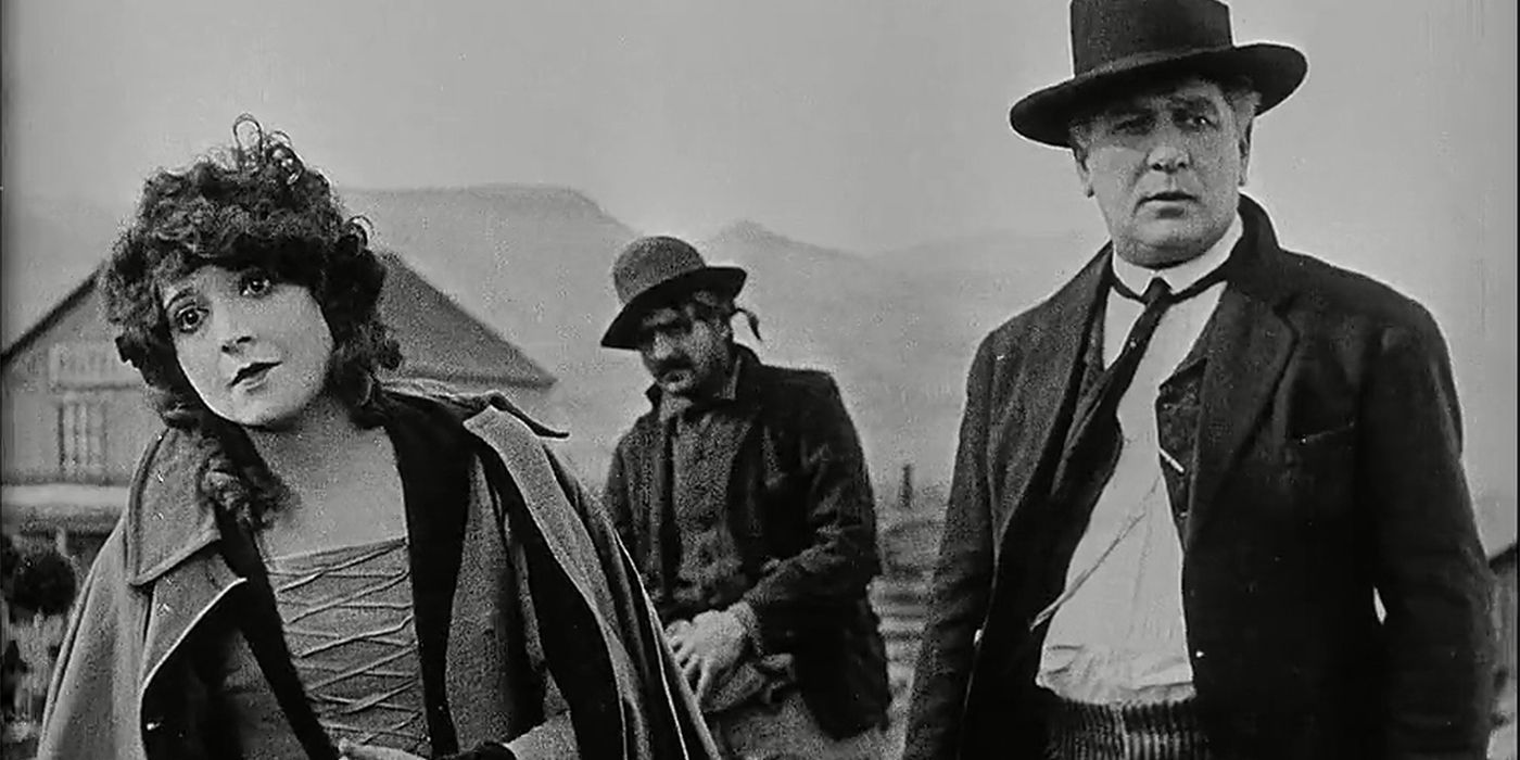 Une femme regarde sur le côté alors que deux hommes se tiennent à côté d'elle en costume occidental dans une scène de The Iron Horse de John Ford.