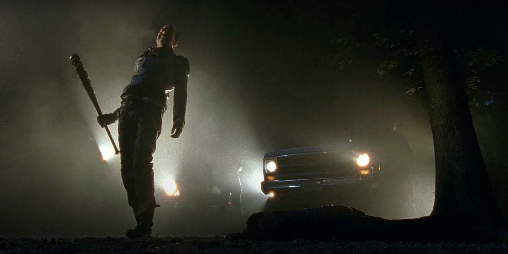 Negan (Jeffrey Dean Morgan) stands delighted before Glenn's (Steven Yeun) body as car headlights stream from behind him.